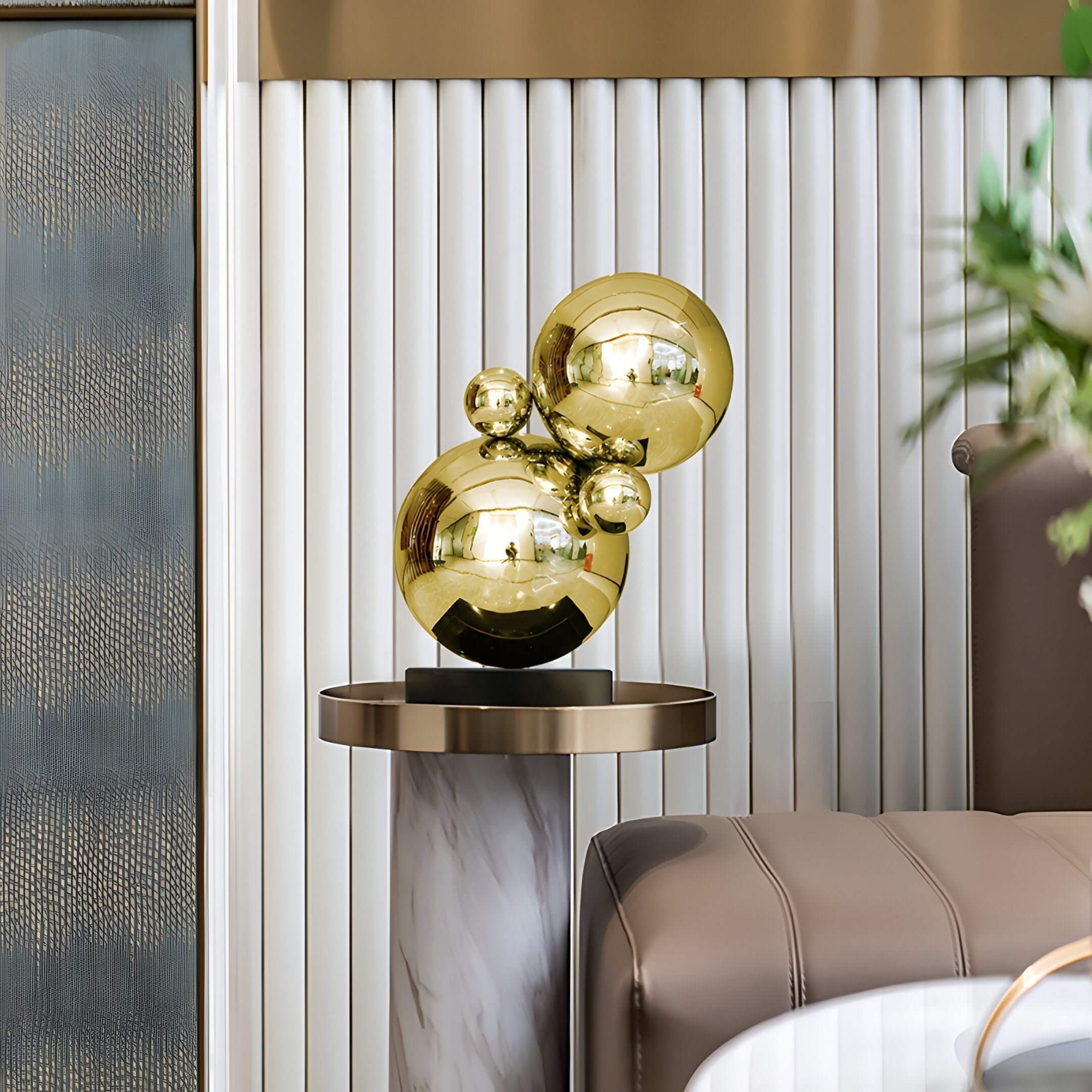 The Molecular II Gold Geometric Steel Sculpture by Giant Sculptures, 55cm, features five interconnected gold spheres on a marble-finished round pedestal. The background has vertical ridged panels, with a brown sofa partially visible to the right.