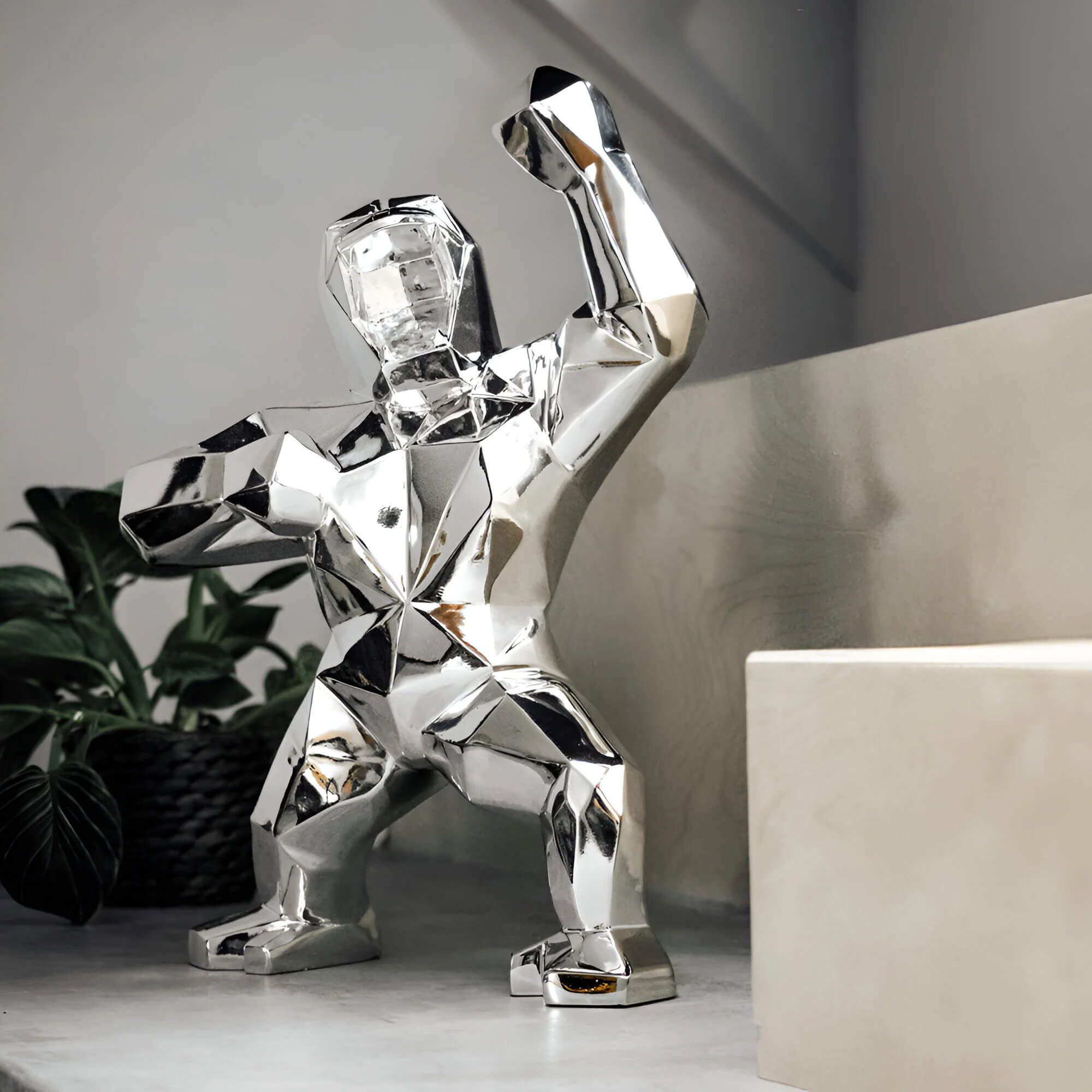 Polished silver geometric gorilla sculpture with a raised fist displayed on a modern shelf, with a potted plant and neutral walls.