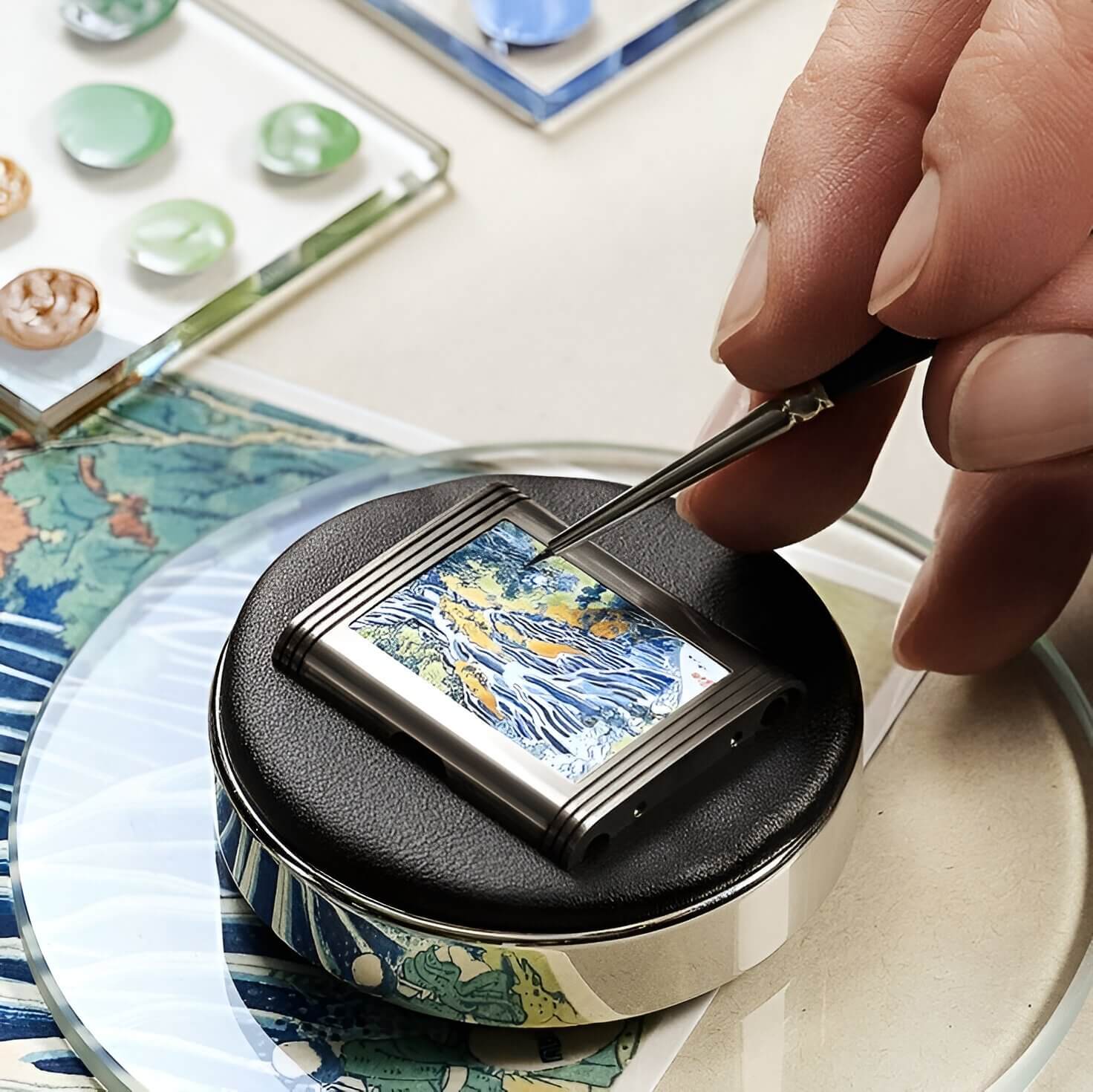 Close-up of a craftsman hand-painting a tiny enamel artwork on a watch case, with colourful enamel samples and tools in the background.