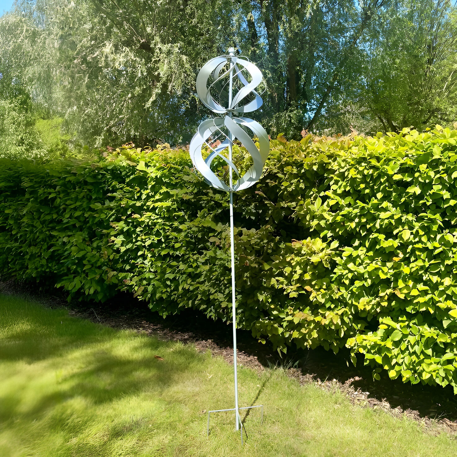 A kinetic garden sculpture featuring dual interlocking metallic spirals on a tall stand, displayed outdoors on green grass with lush hedges in the background.