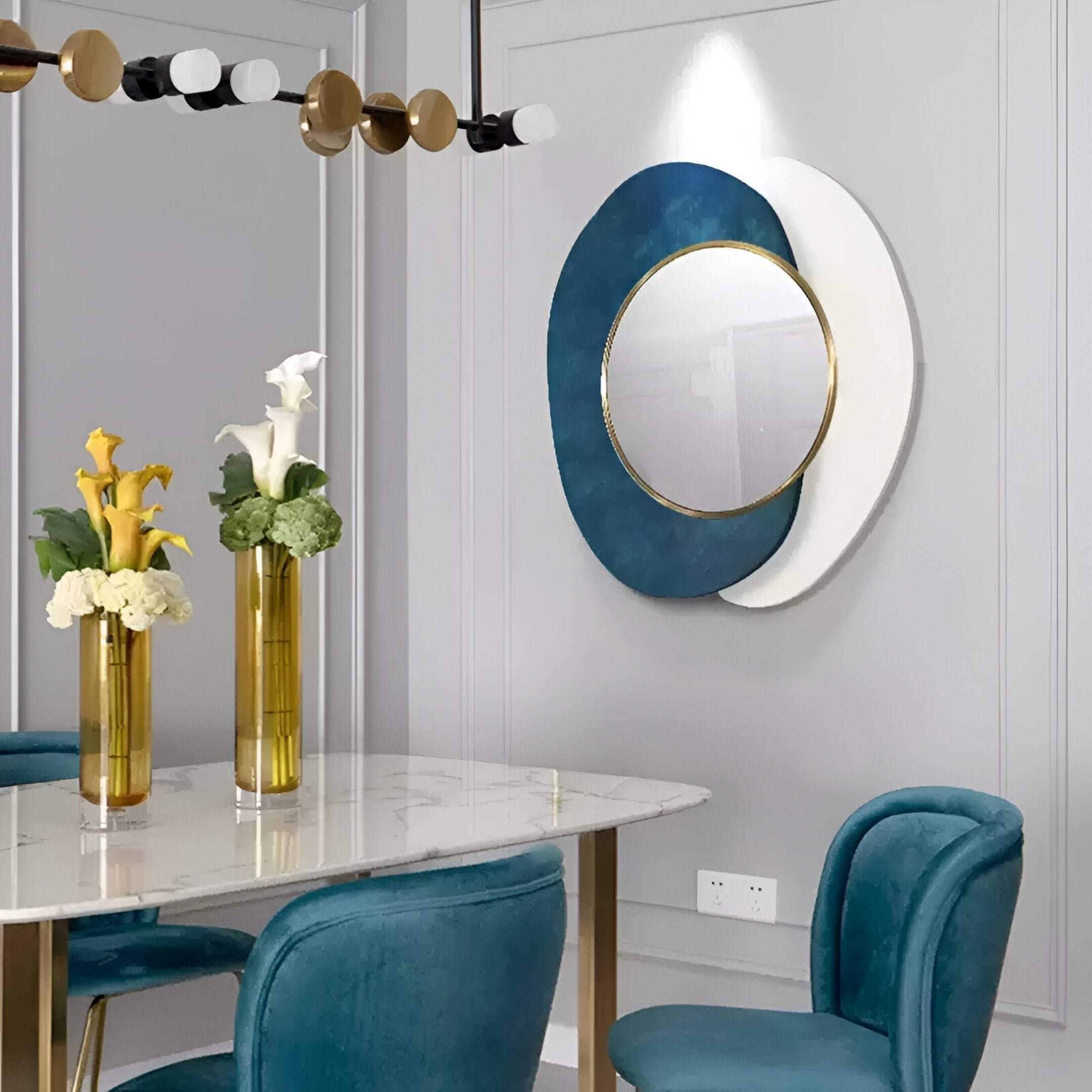 A modern dining room with a circular blue and white abstract wall mirror framed in gold, styled with teal velvet chairs and a marble-top dining table.