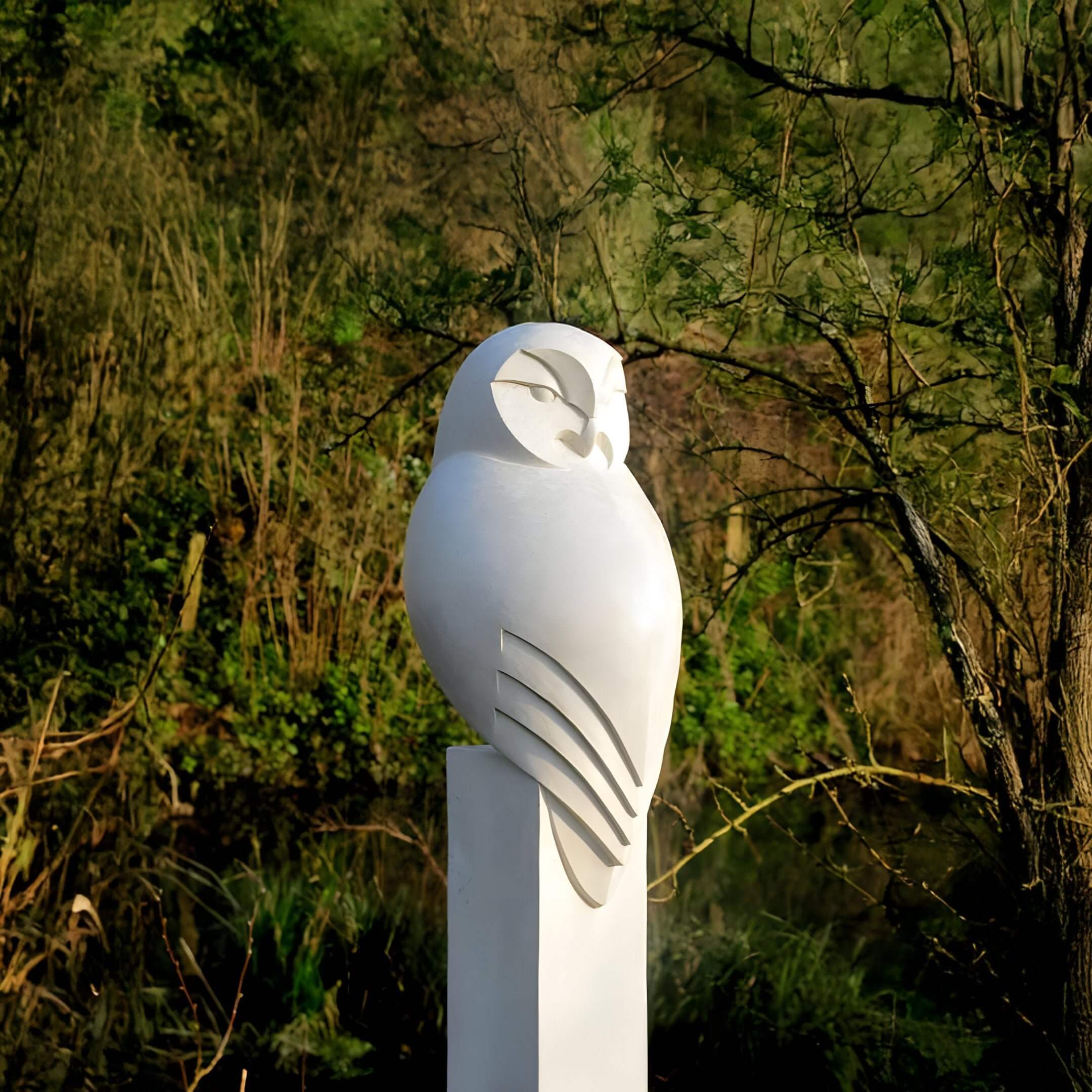 The Giant Sculptures Minimalist Perched Owl Marble Outdoor Sculpture - 40cm, featuring a smooth white marble owl on a pedestal, elegantly contrasts against lush green foliage and trees in any natural setting.