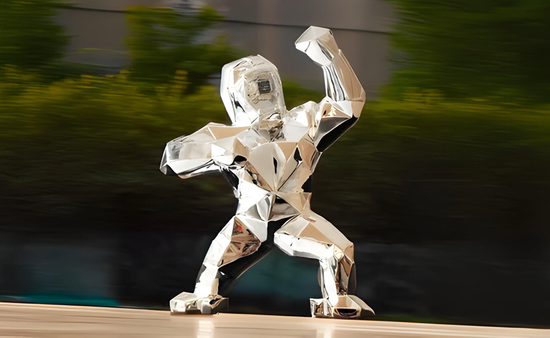 Polished silver geometric gorilla sculpture with raised fists, displayed outdoors with blurred green foliage in the background.