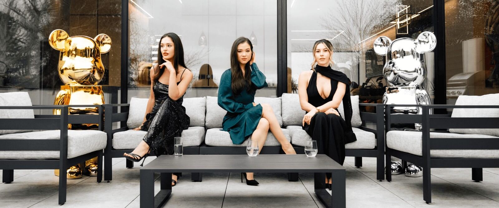 Three women seated outdoors on modern patio furniture, flanked by large gold and silver bear sculptures.