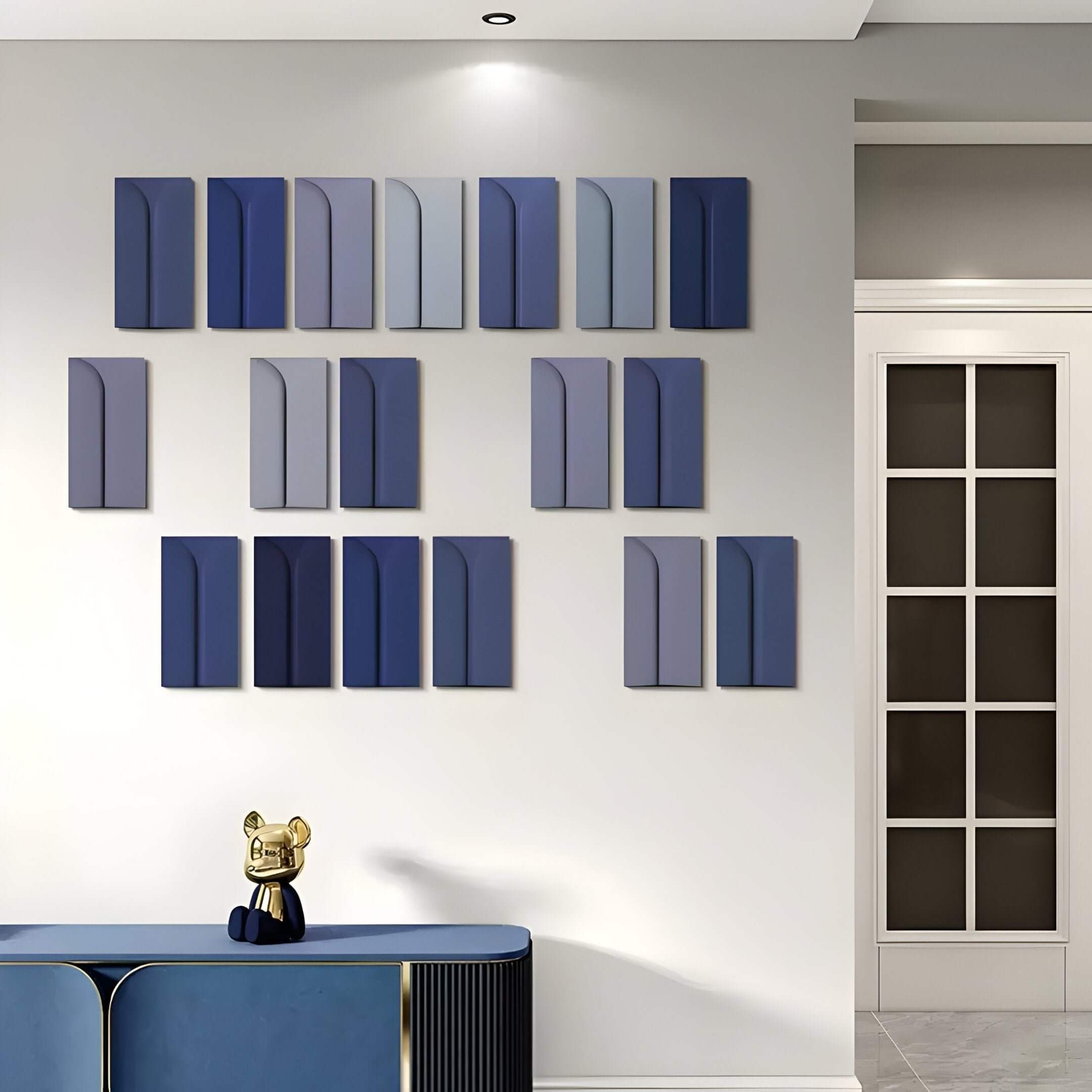 A modern, Nordic minimalistic space showcases the Eximia Blue Tones Multi-Panel Wood Relief 3D Wall Art by Giant Sculptures on a light wall. Below, a sleek blue console table bears a small golden cat figurine. A door with glass panels to the right completes this bright, minimalistic setting.