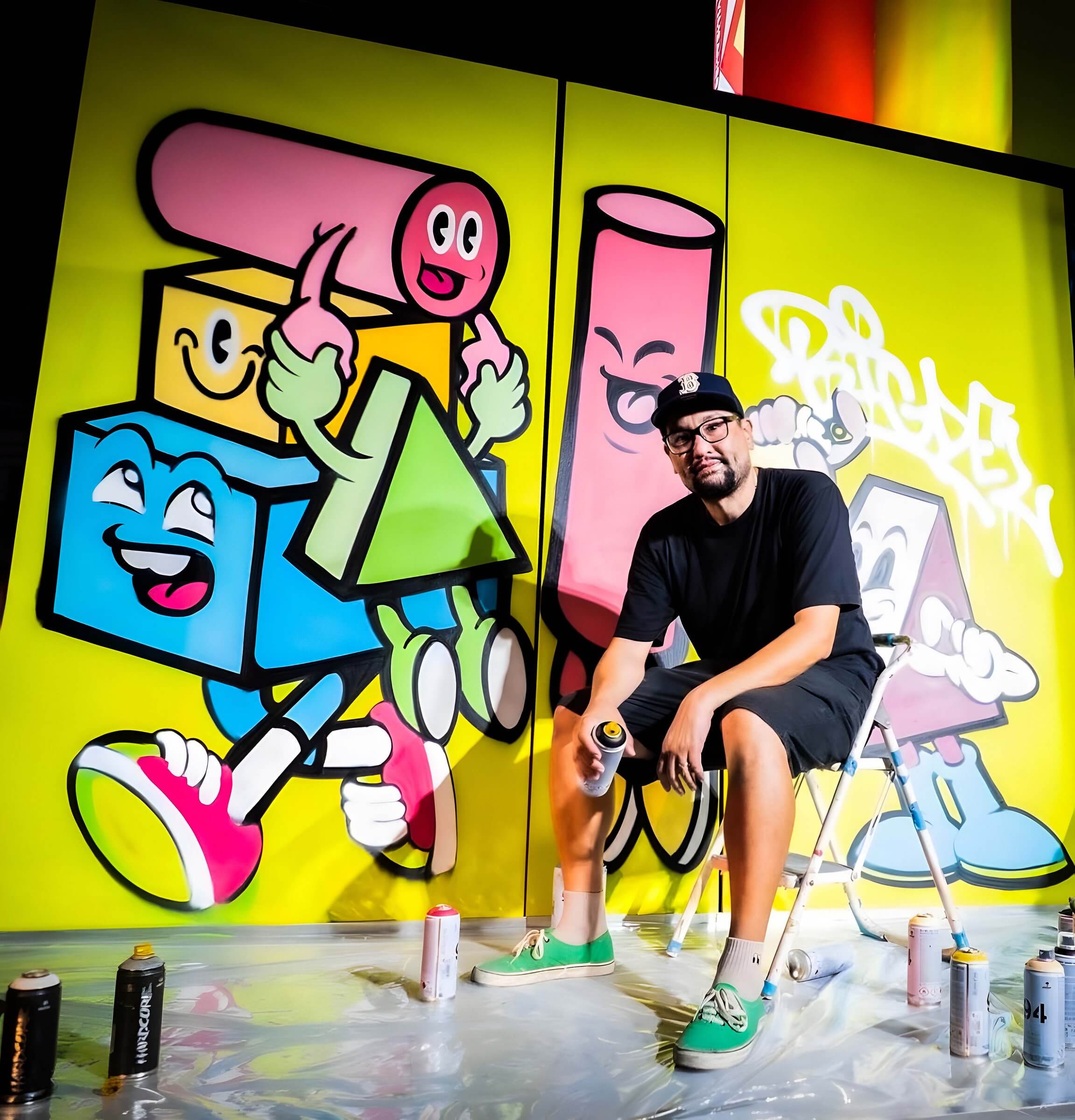 Artist signs an artwork card by hand and then poses in sneakers beside vibrant geometric graffiti characters on a large mural
