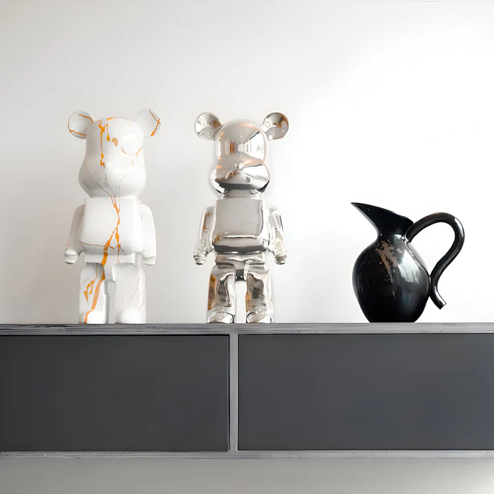 Two bear-shaped sculptures a white one with gold marbling and a reflective chrome one—stand on a minimalist shelf beside a black ceramic pitcher against a white wall.