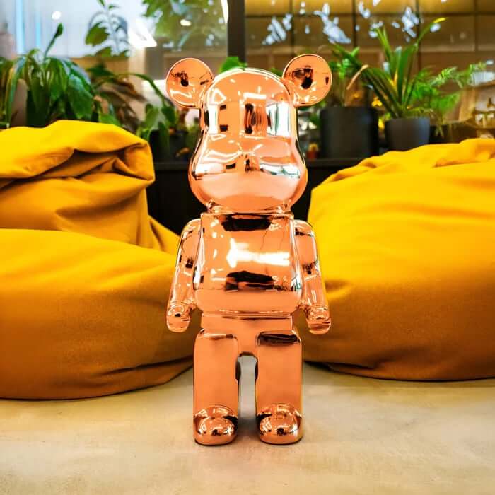 Chrome rose-gold resin bear sculpture between mustard-yellow beanbags with indoor plants in the background.
