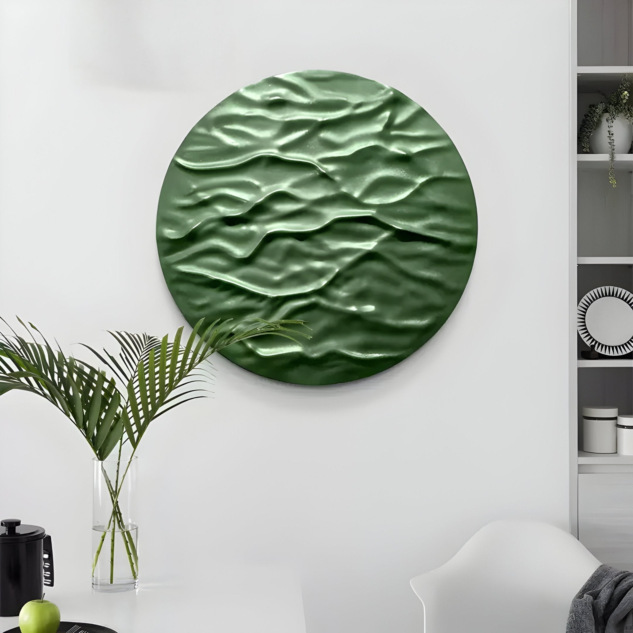 A modern interior features the Orenda Ripple Wave Green Wood Relief 3D Wall Art by Giant Sculptures, with its wavy pattern. Below, a table with a glass vase of palm leaves rests against minimalist white walls and shelves, completing the decor.