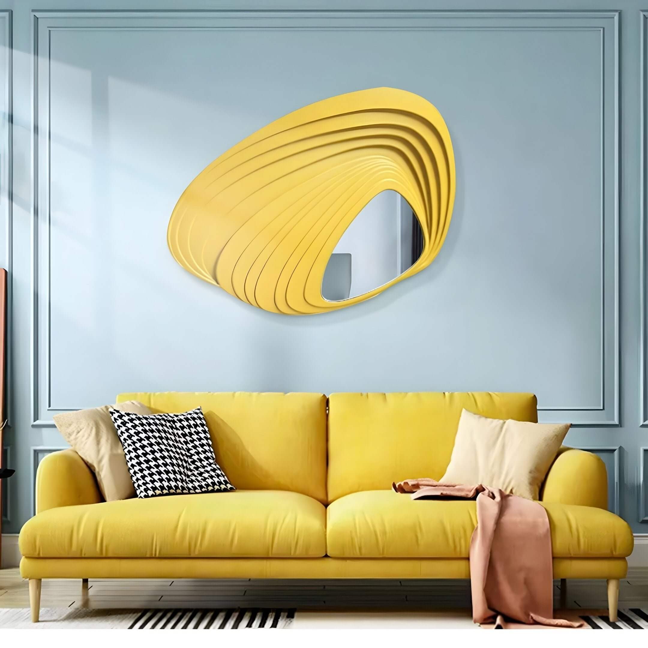 In a contemporary living room, a yellow couch decorated with houndstooth and plain pillows and a soft pink blanket is complemented by the Reflecta Yellow Wood Carving Mirror 3D Wall Art from Giant Sculptures, adding modern flair against light blue walls.