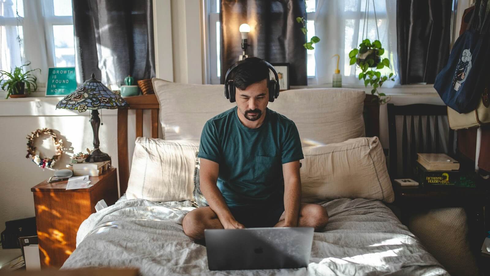 A man sits on a bed with his laptop and headphones on
