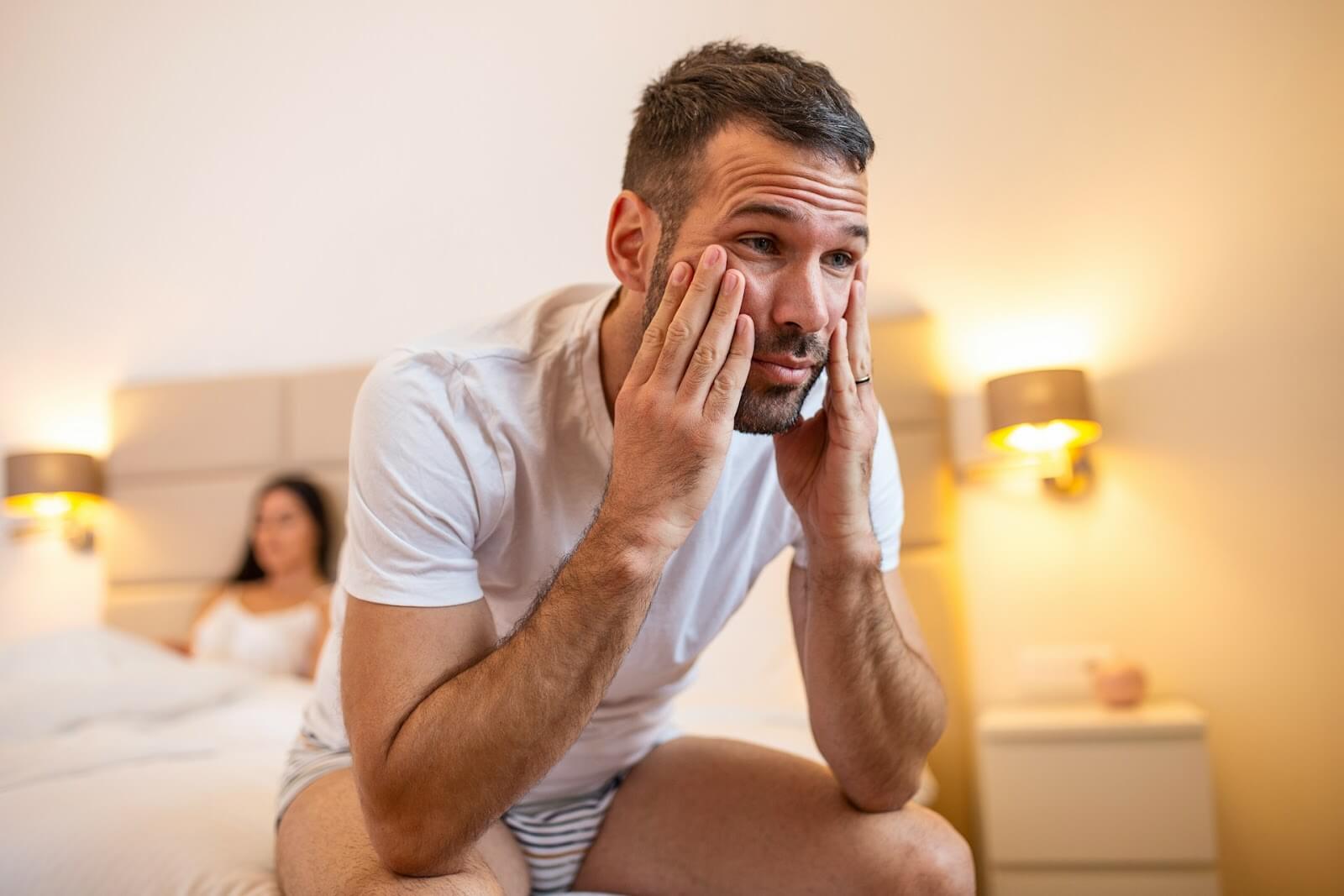 A man looks sad on the edge of his bed, his wife behind him, suffering from a lack in sexual performance