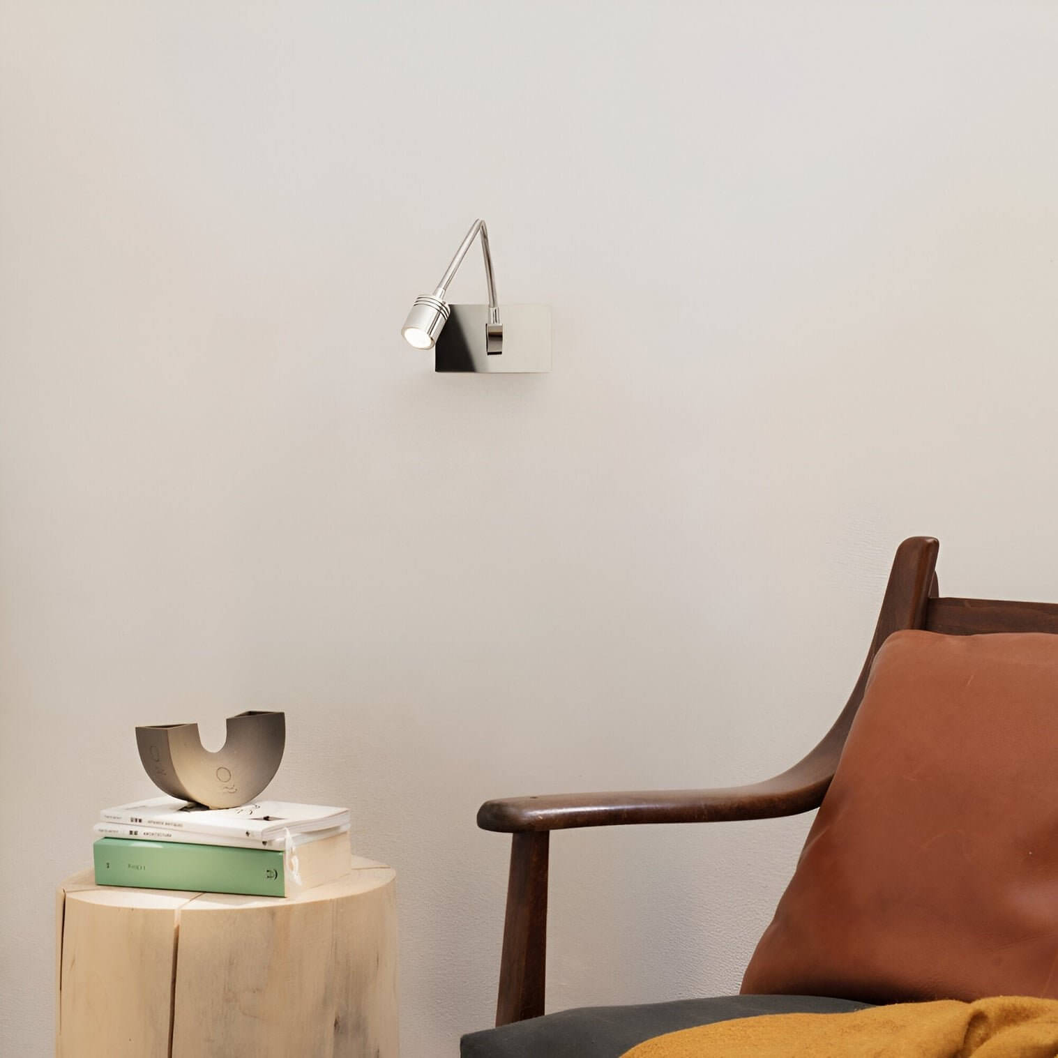 Minimalist reading nook with chrome adjustable wall lamp, wooden chair with brown cushion, and round wooden side table with stacked books.