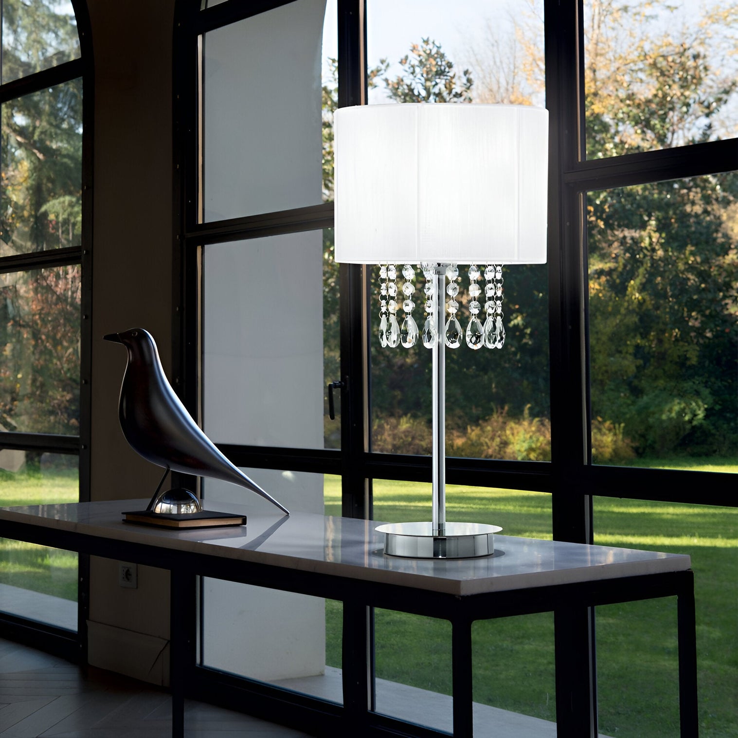 Chrome table lamp with white shade and hanging crystals beside a sculptural black bird.