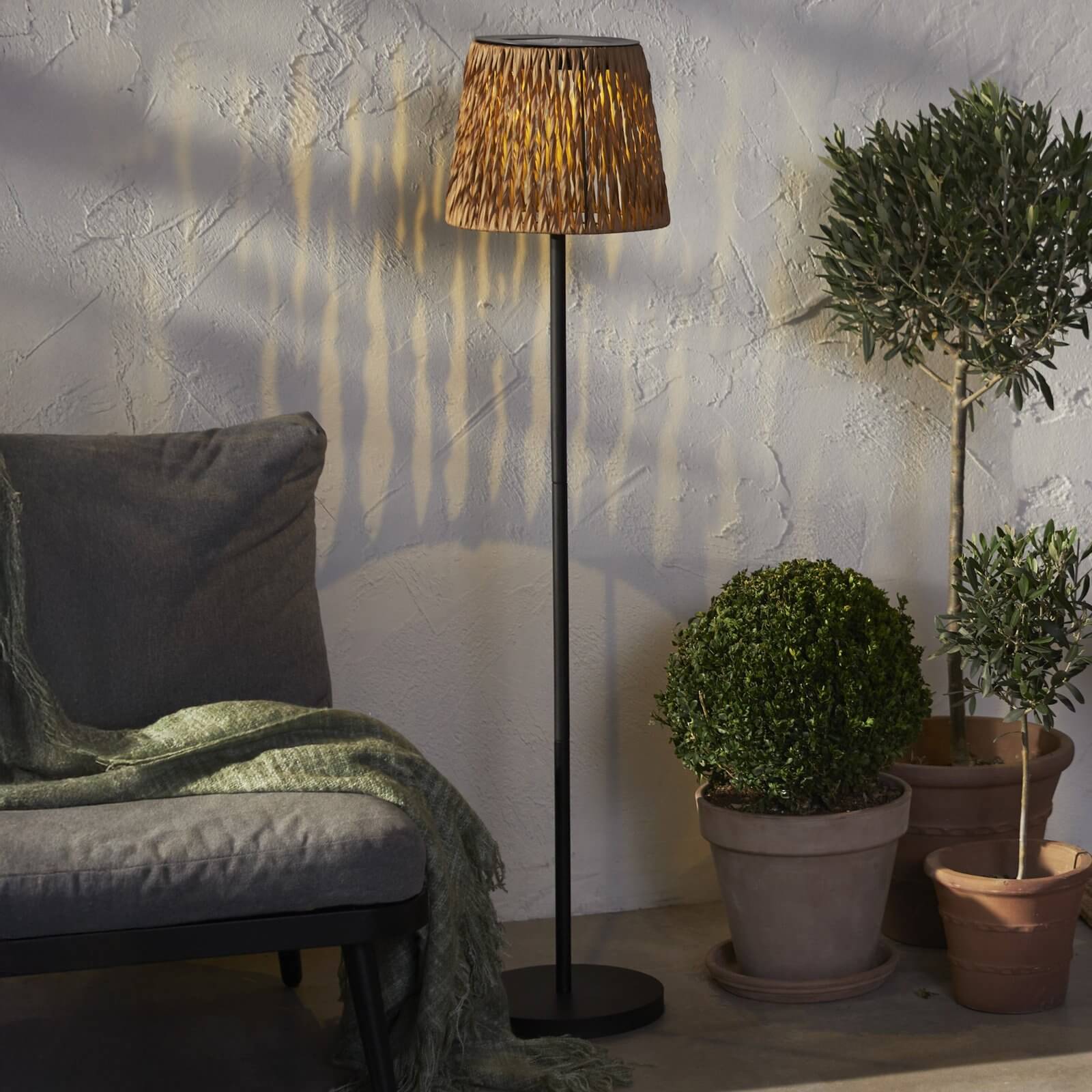 Floor lamp with woven rattan shade casting shadow patterns beside grey seating and potted shrubs on a patio.
