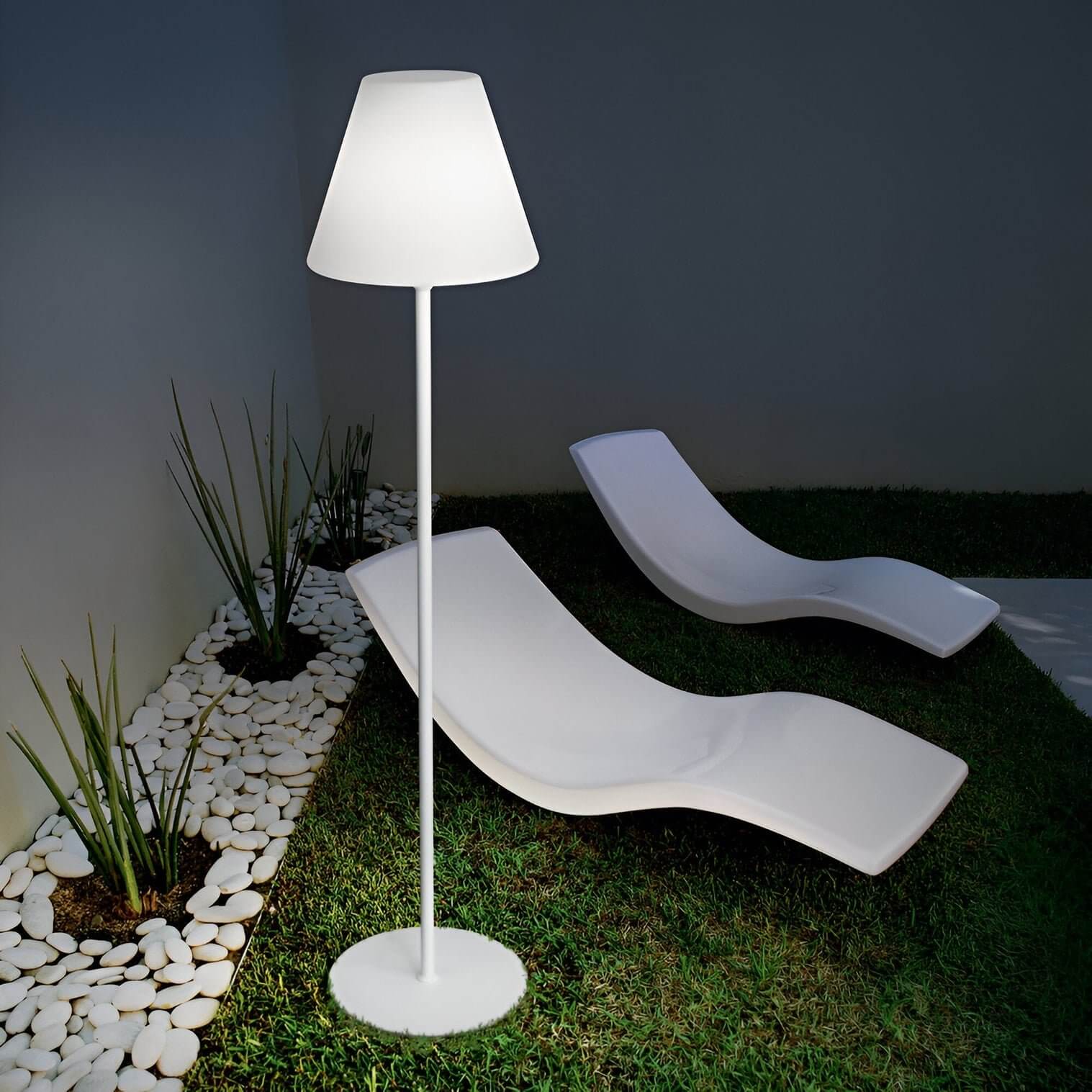White floor lamp with a cone-shaped shade placed on grass beside two modern white sun loungers and white stones.