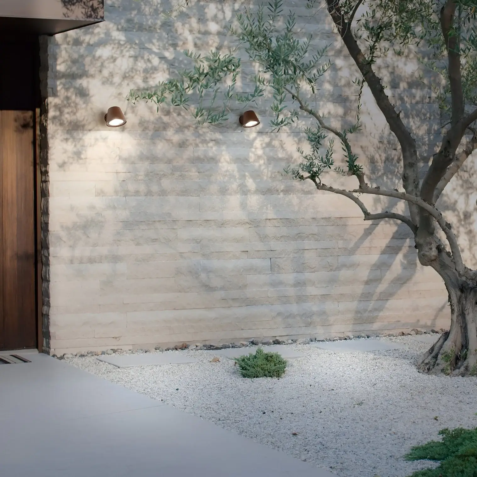 Minimalist home entrance with two small brown wall-mounted lights casting soft glow on textured stone wall beside an olive tree.