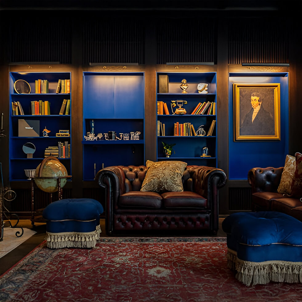 A classic library with blue shelving and gold picture lights illuminating books and decor. The space features a leather Chesterfield sofa, tufted ottomans, and a Persian rug.