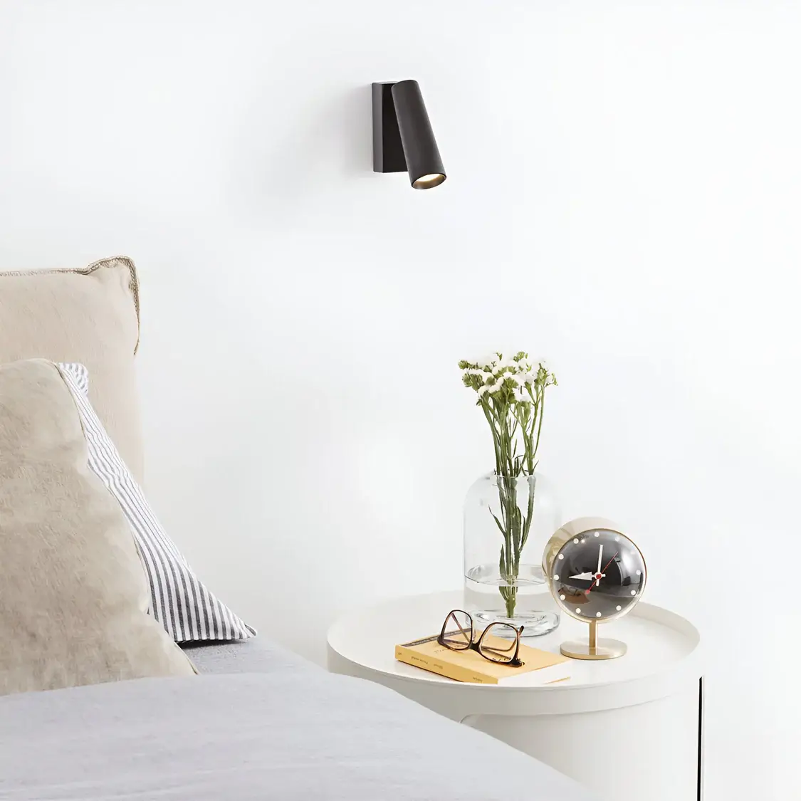 A minimal bedside setup with a sleek black wall-mounted sconce providing focused lighting. The side table holds a modern clock, glasses, and a vase with fresh white flowers, complementing the neutral tones of the bed.