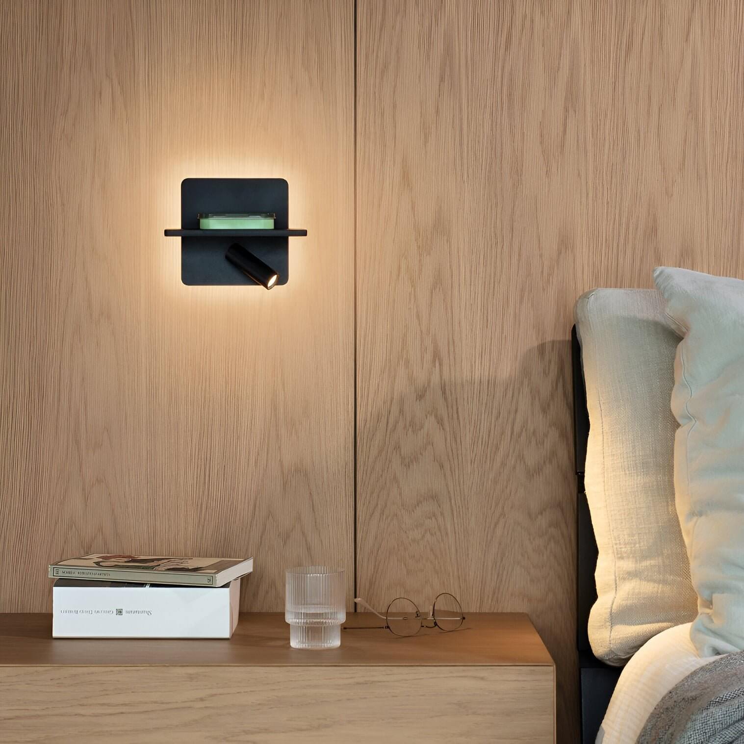 Contemporary bedroom with wooden wall panels, black wall lamp with integrated shelf, and bedside table with books, glass, and eyeglasses.