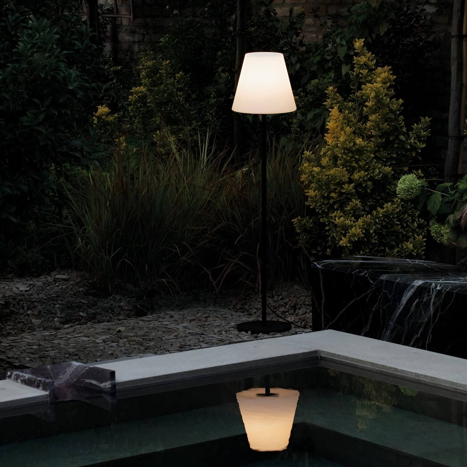 Tall floor lamp with classic white shade reflecting in a pool surrounded by dense garden foliage at night.