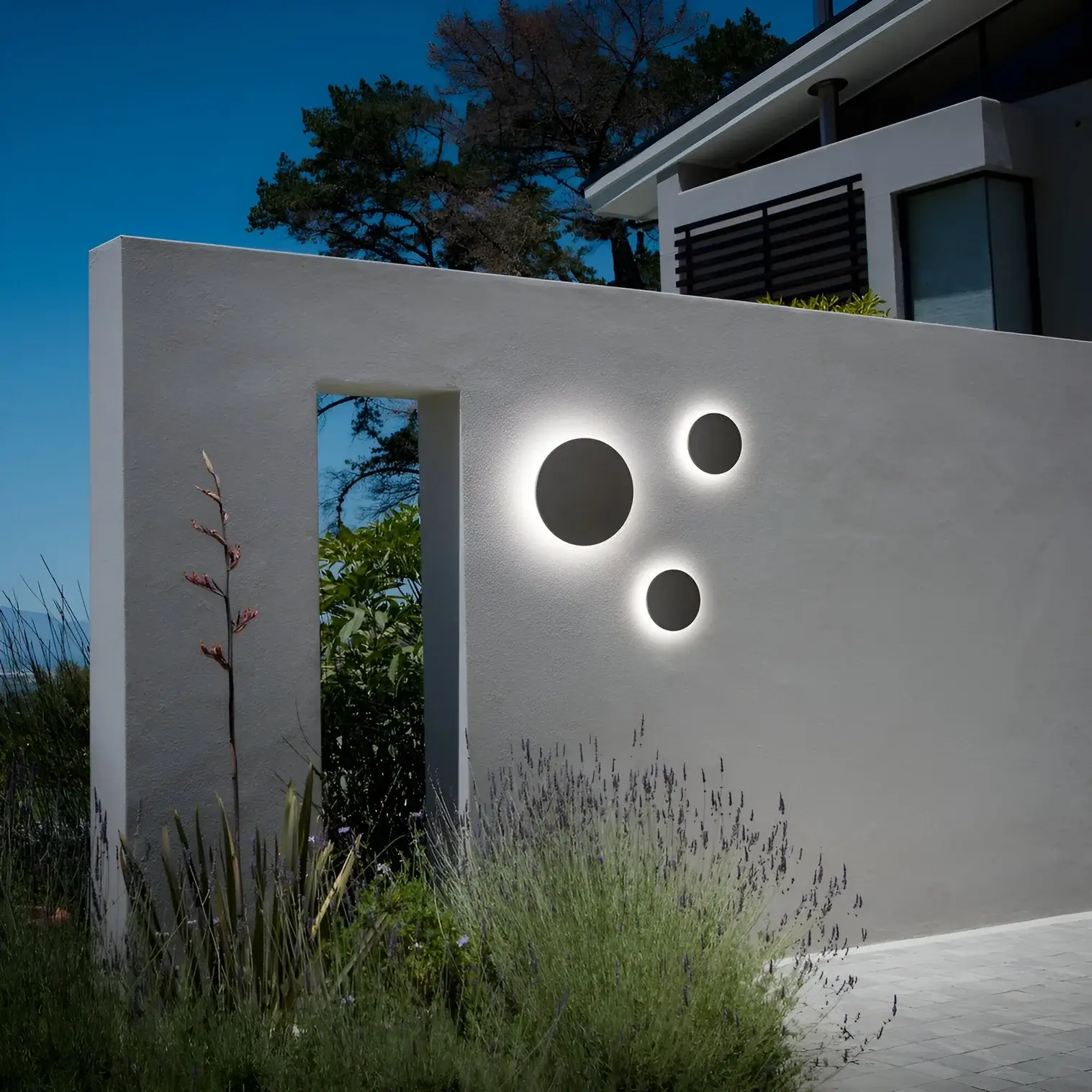 White exterior wall with circular recessed lights glowing at night, surrounded by lavender and tall grasses.