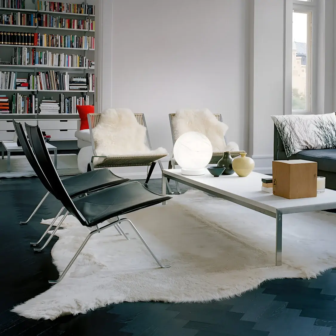 A sleek living space with black leather chairs, a white coffee table adorned with ceramic vases and a glowing globe lamp, surrounded by a built-in bookshelf and accented with a soft white rug on dark flooring.