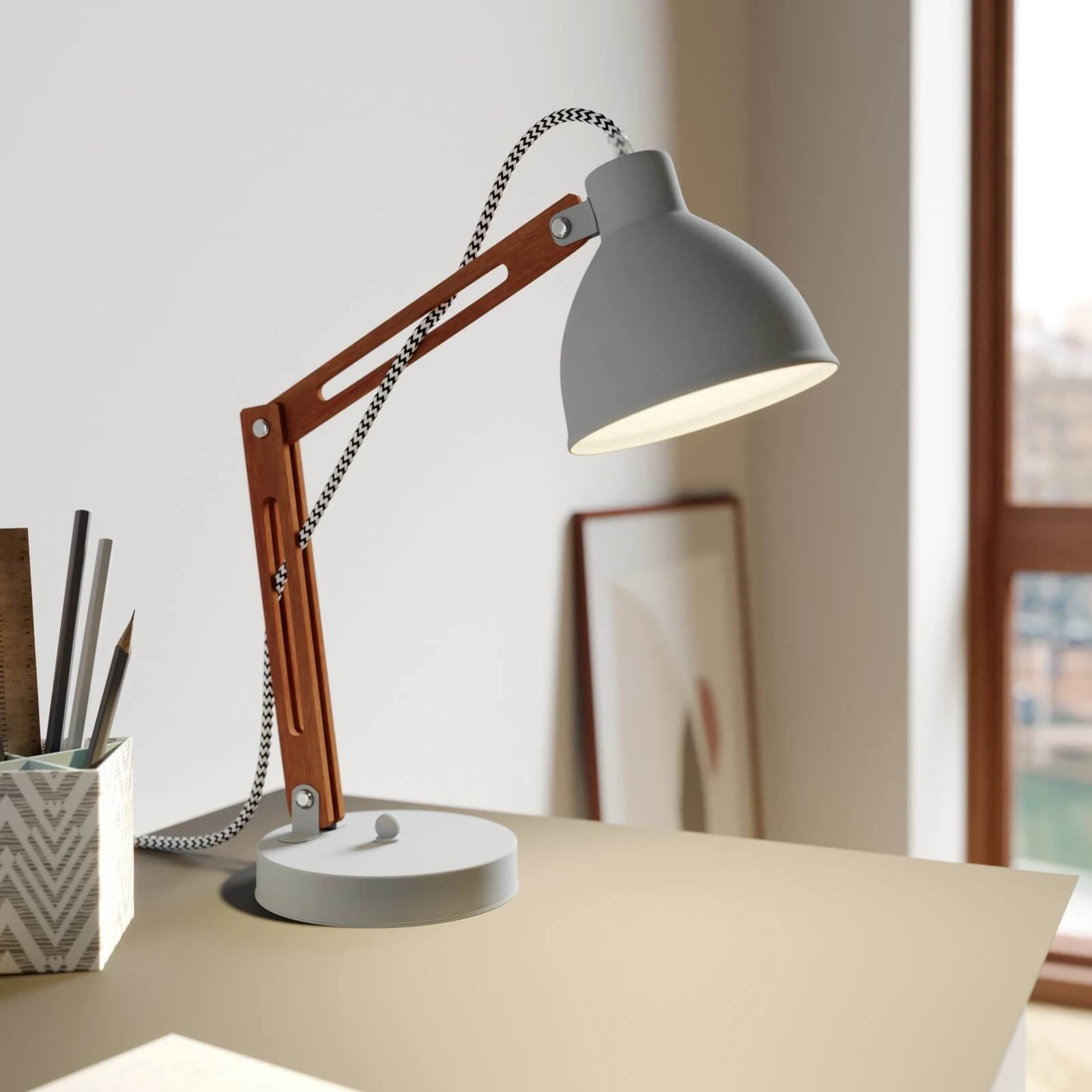 Grey and wood desk lamp with exposed cord on a tidy work surface in front of a sunlit window.