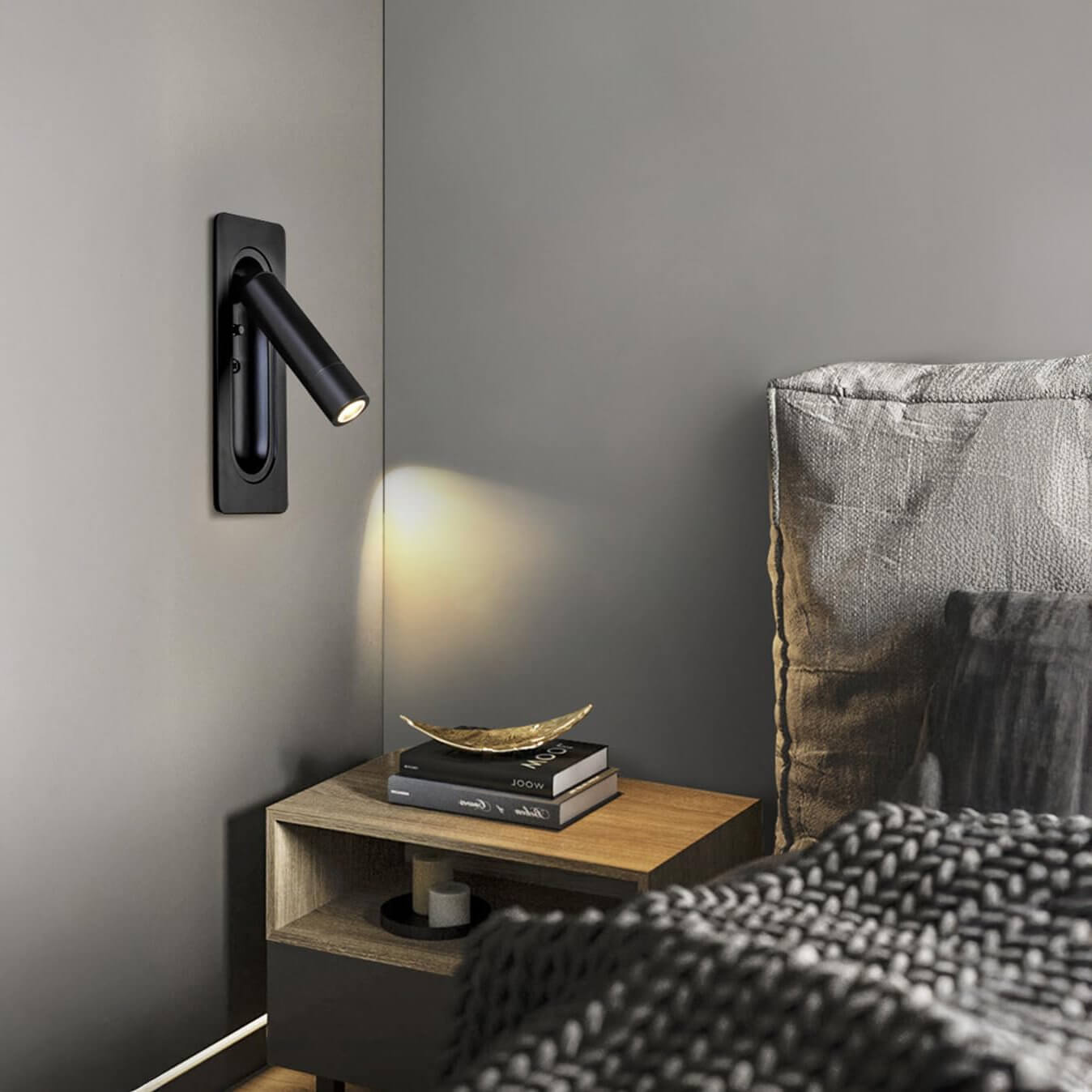Bedroom with matte black cylindrical wall lamp, grey wall, wooden bedside table, and layered black-and-gold decorative accents.