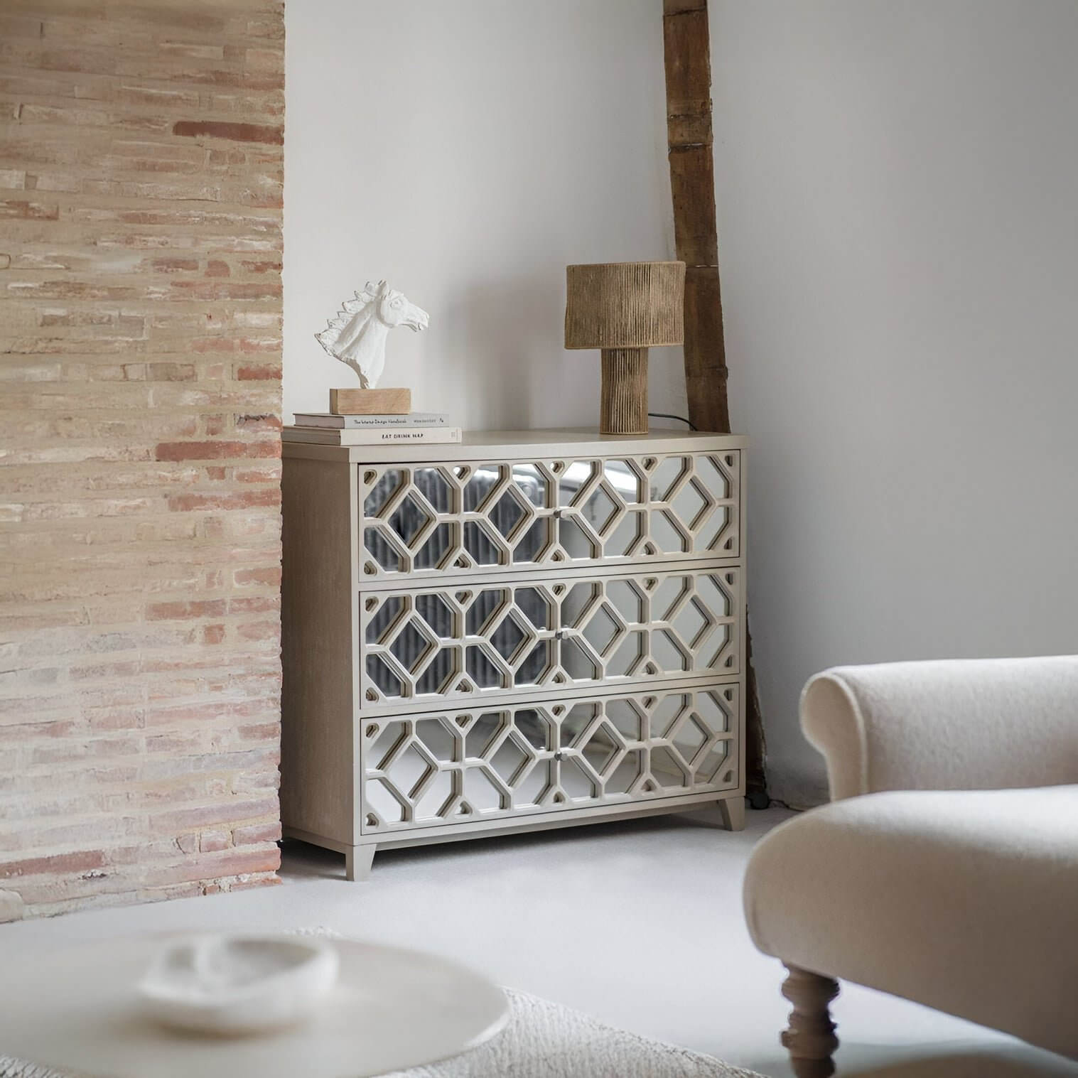 Wood block lamp on mirrored geometric dresser beside cream upholstered chair & exposed brick