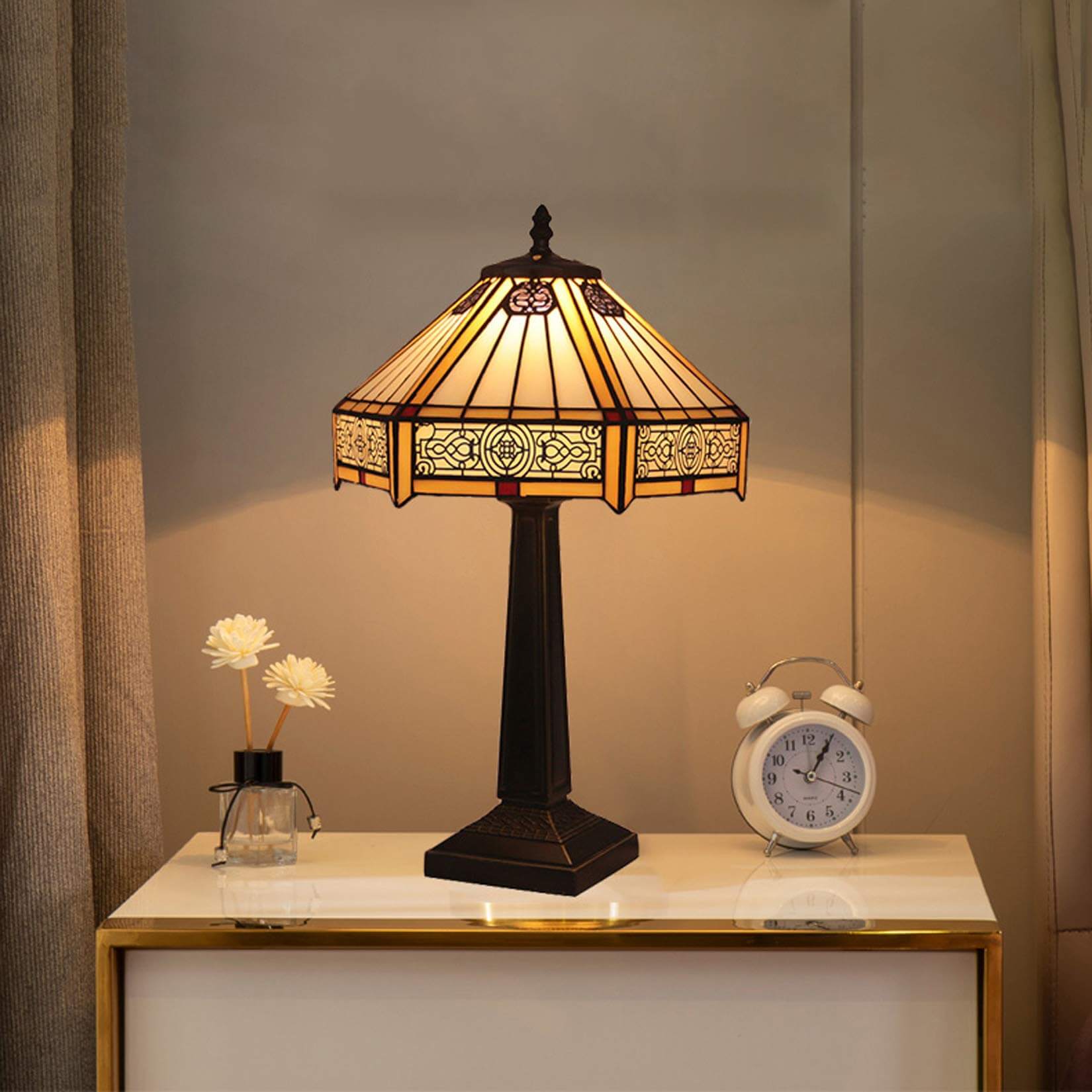 Classic Tiffany-style table lamp with ornate shade on a nightstand beside an alarm clock and decorative flowers
