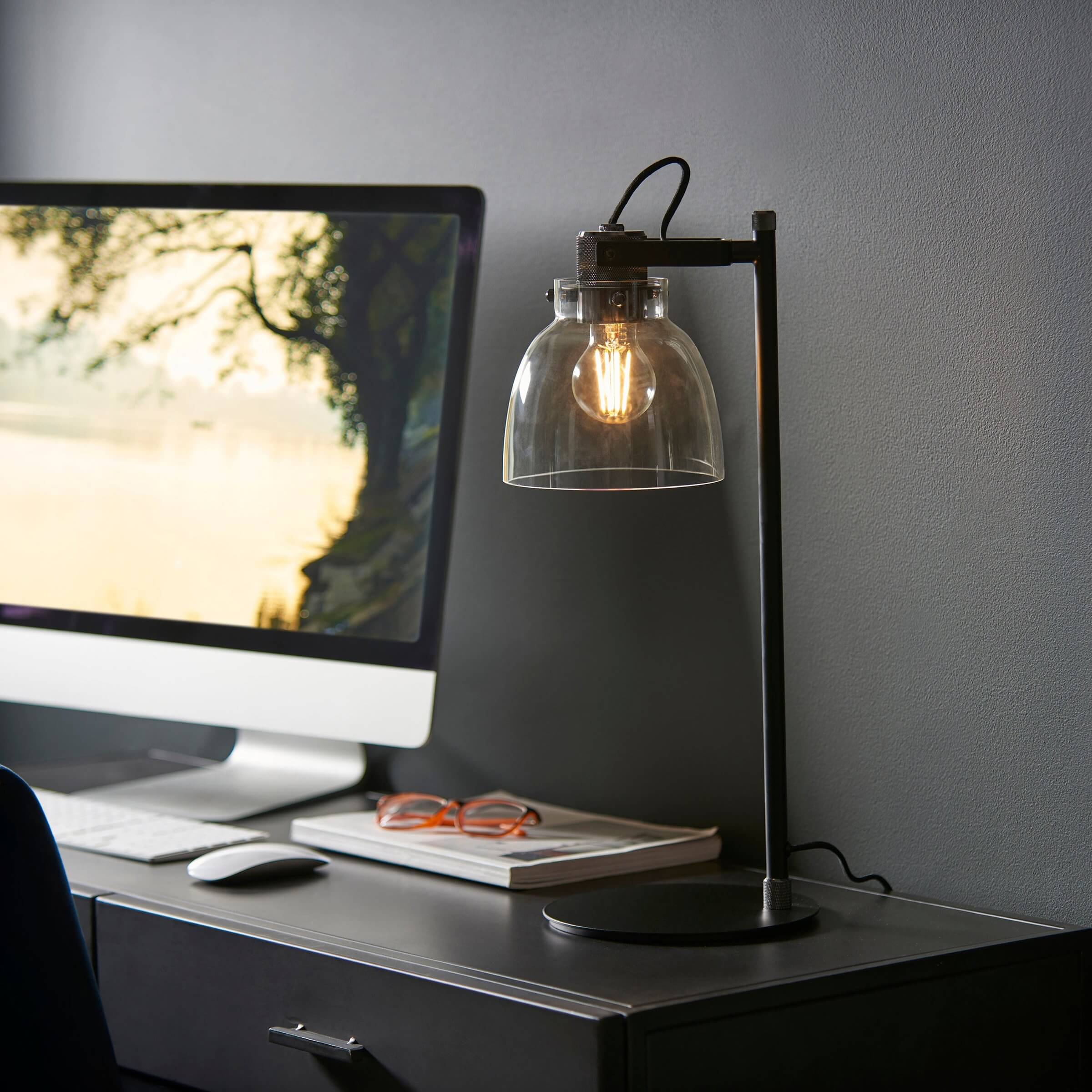 Industrial table lamp with clear glass dome shade and exposed filament bulb on a black desk beside a computer.