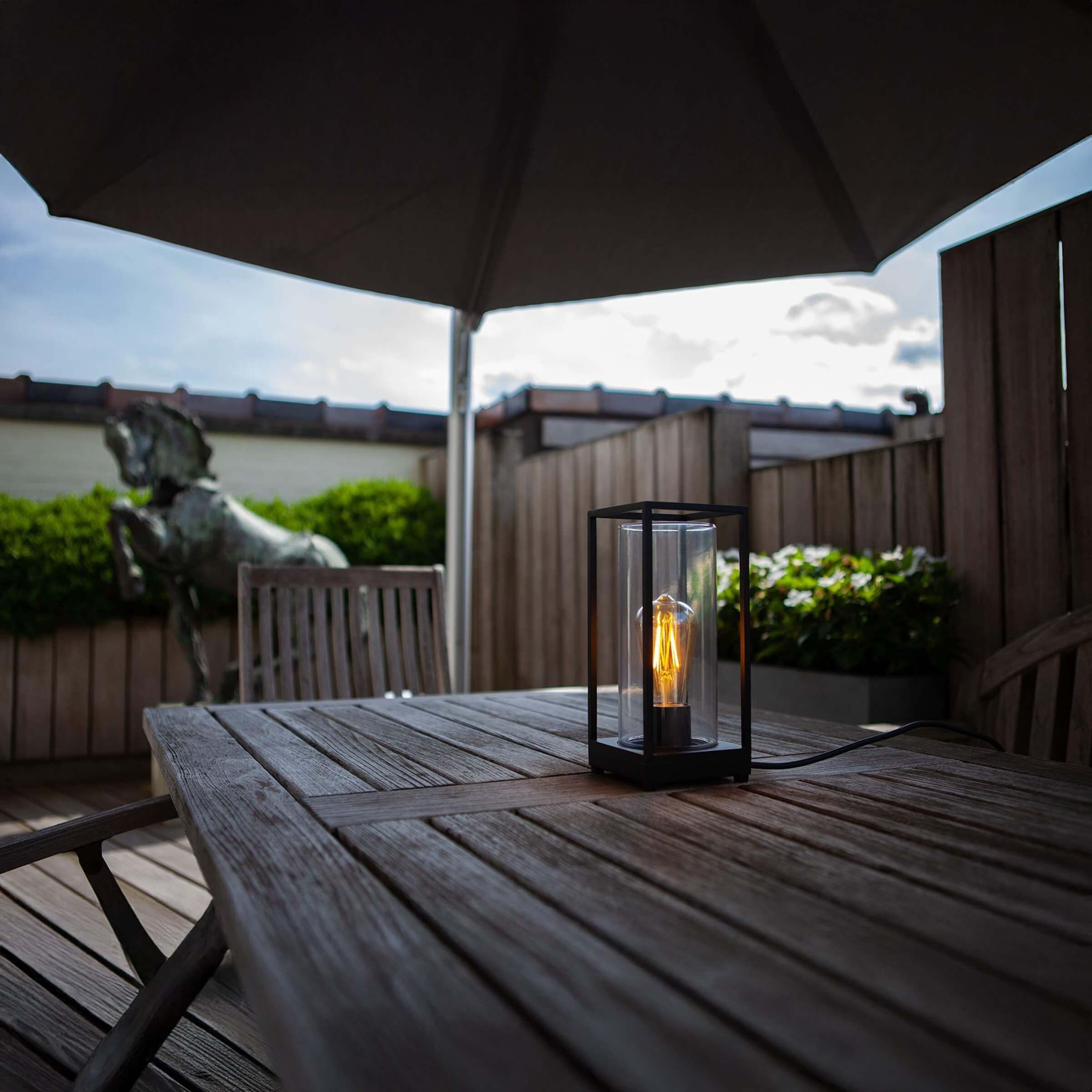Black metal lantern table lamp with exposed Edison bulb on a wooden patio table.