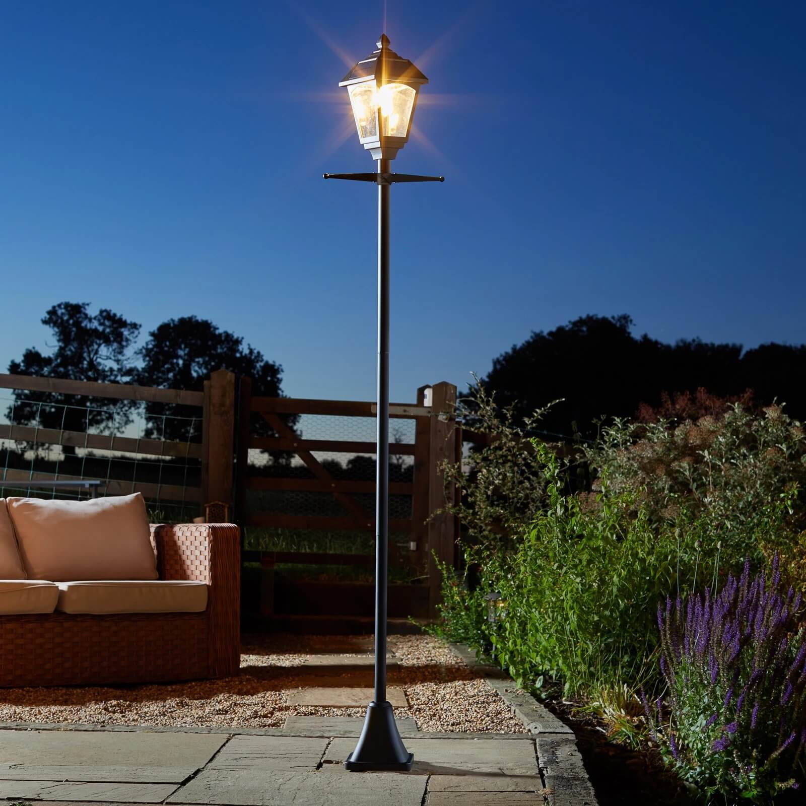 Classic outdoor lantern-style lamp post lit at night on stone patio with garden seating