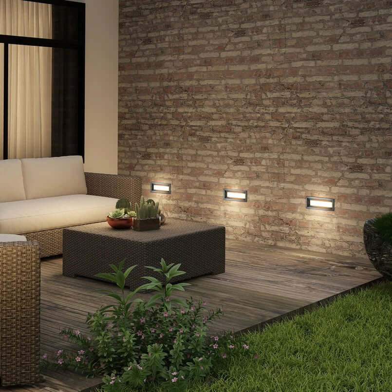 Brick wall with three rectangular recessed lights illuminating a patio seating area with wicker furniture and plants.