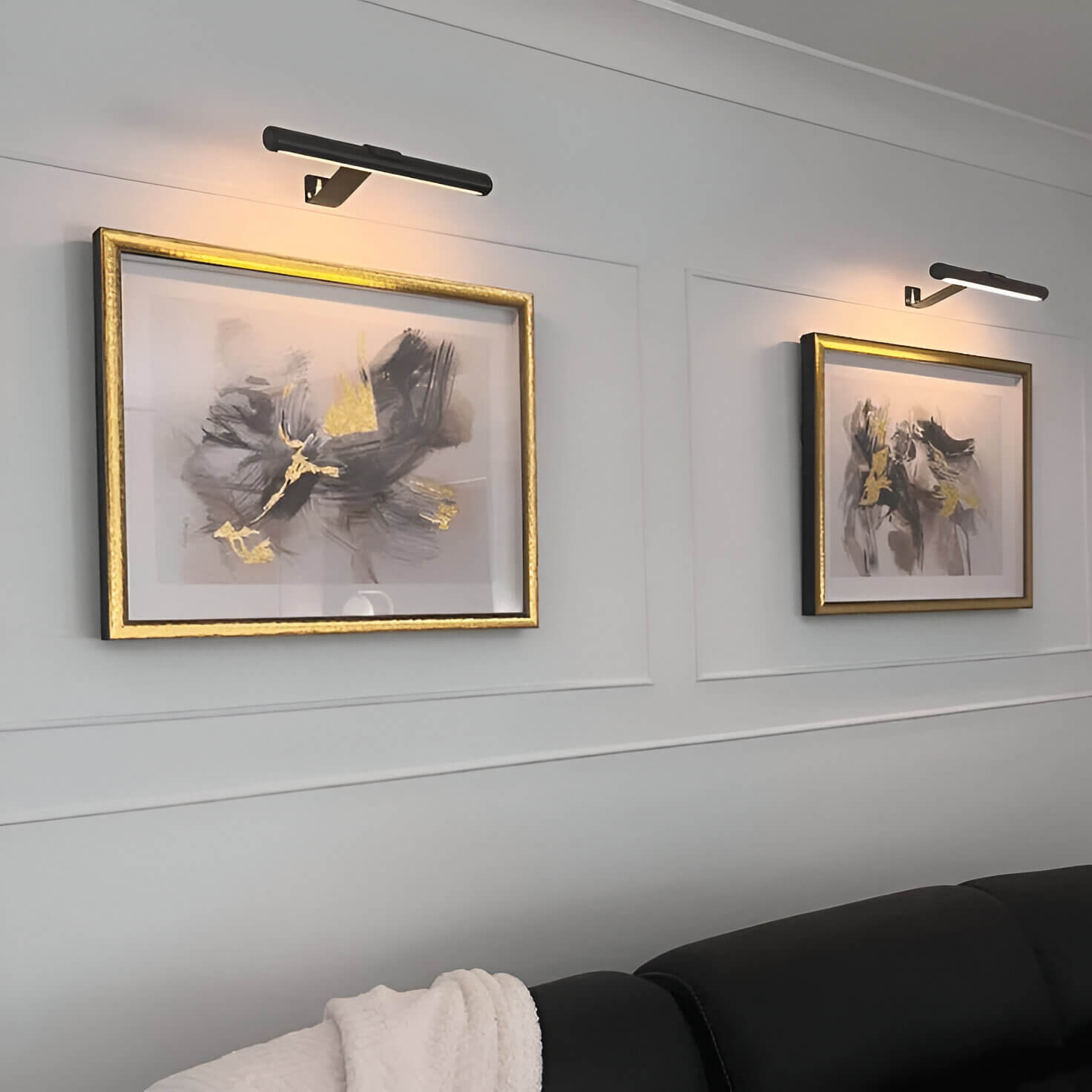  Two contemporary picture lights in black finish, casting a warm glow over framed abstract artwork with gold accents, mounted on a clean white wall.