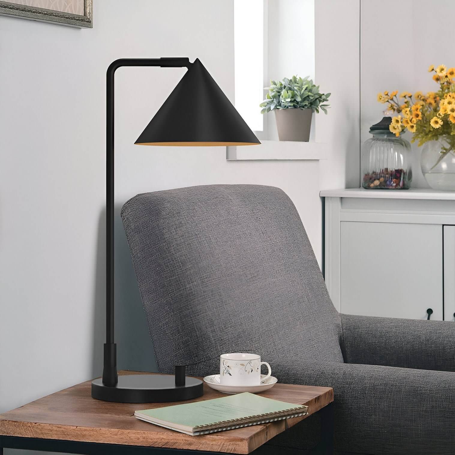 Black table lamp with conical shade and straight arm on a wooden side table next to a grey upholstered armchair.