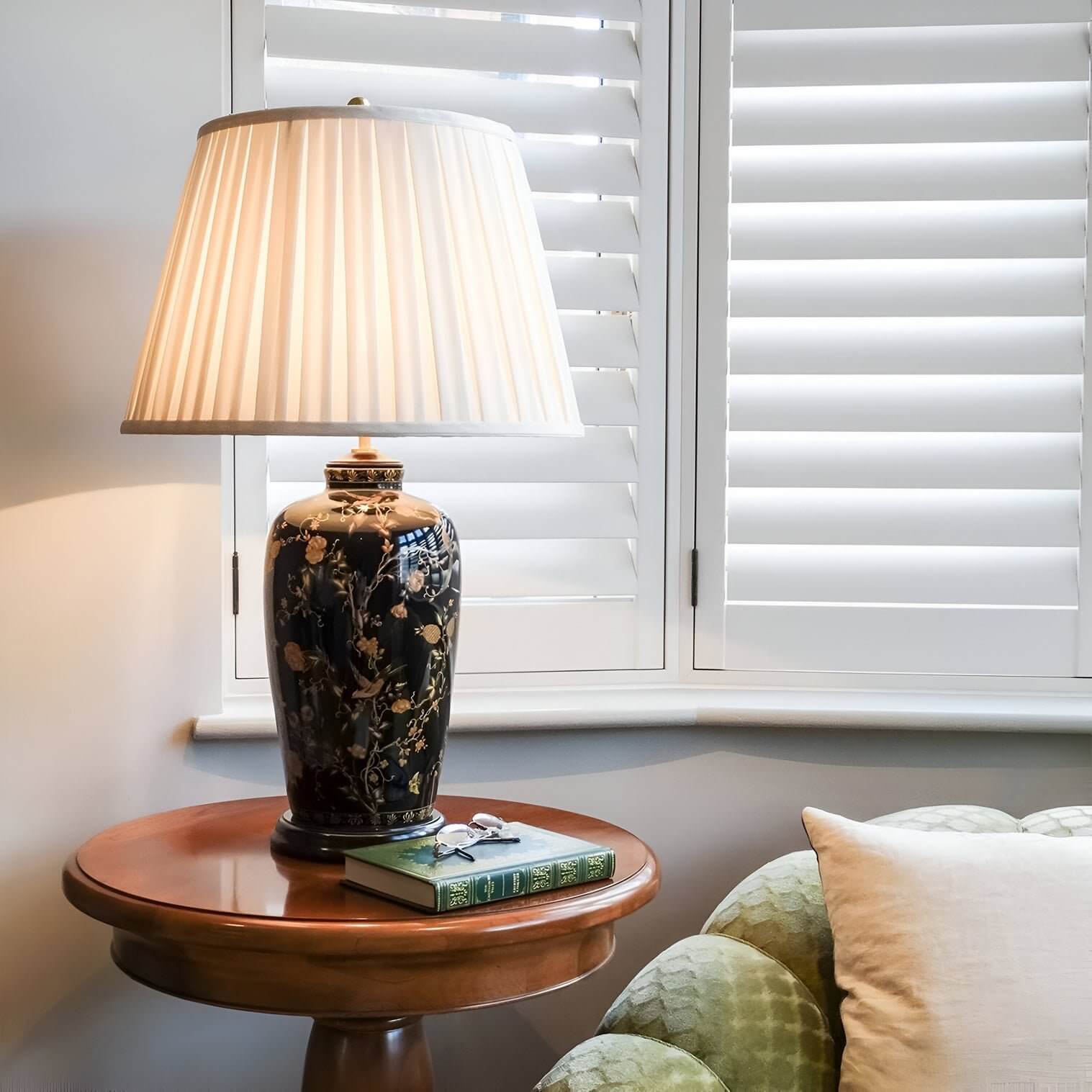 Floral ceramic table lamp with pleated shade on round wood table near shuttered window