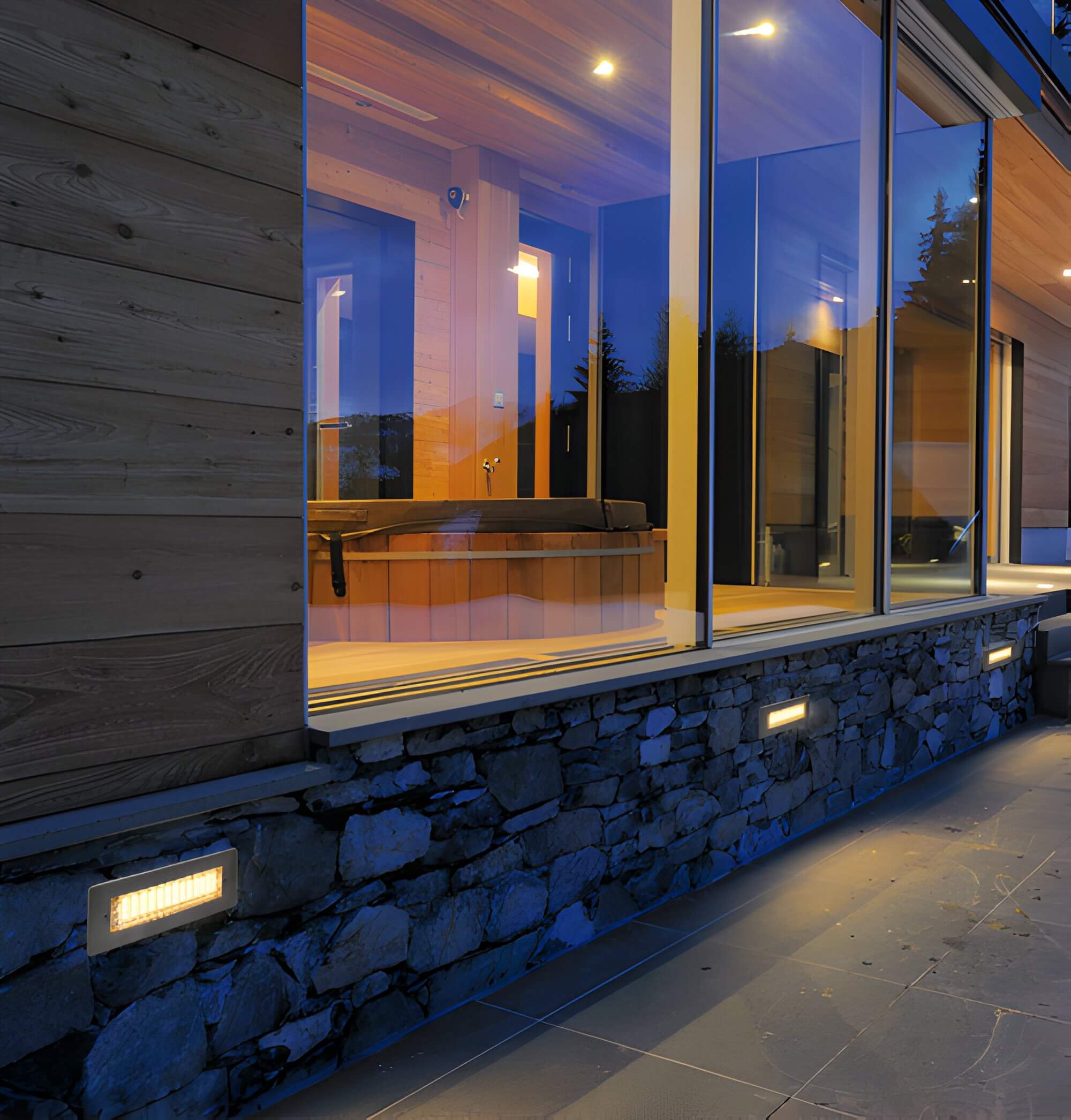 Recessed lights set in a stone wall below large glass windows of a modern home with warm interior lighting.