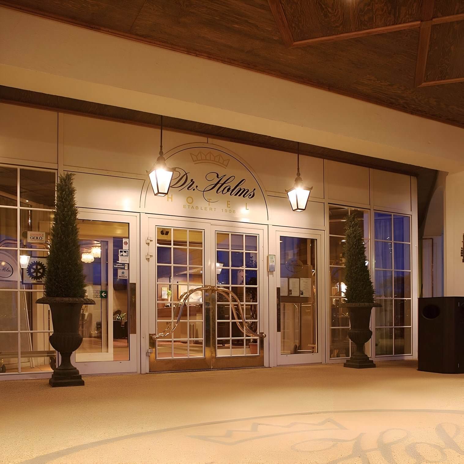 Hotel entrance at dusk with hanging lanterns, double glass doors, and symmetrical topiary planters on either side.
