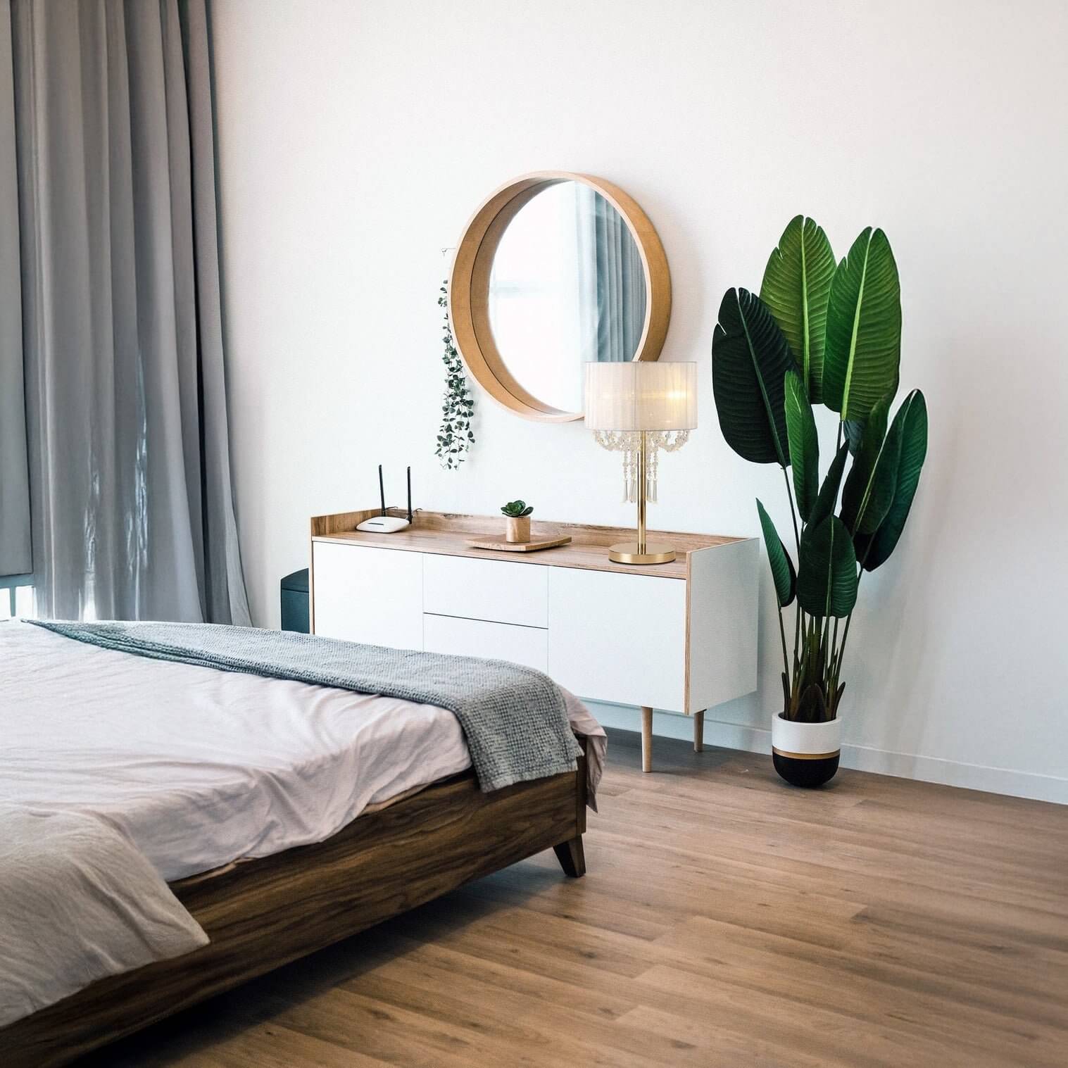 Bright bedroom corner with a wooden dresser, gold-accented table lamp, round wall mirror, and a large indoor plant.