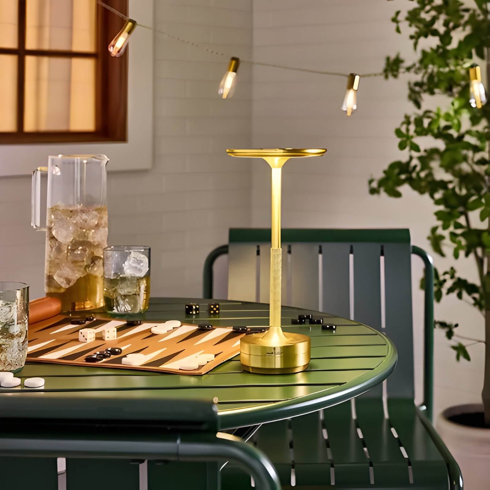 Slim brass outdoor table lamp on a green metal table with backgammon board, ice pitcher, and string lights above.