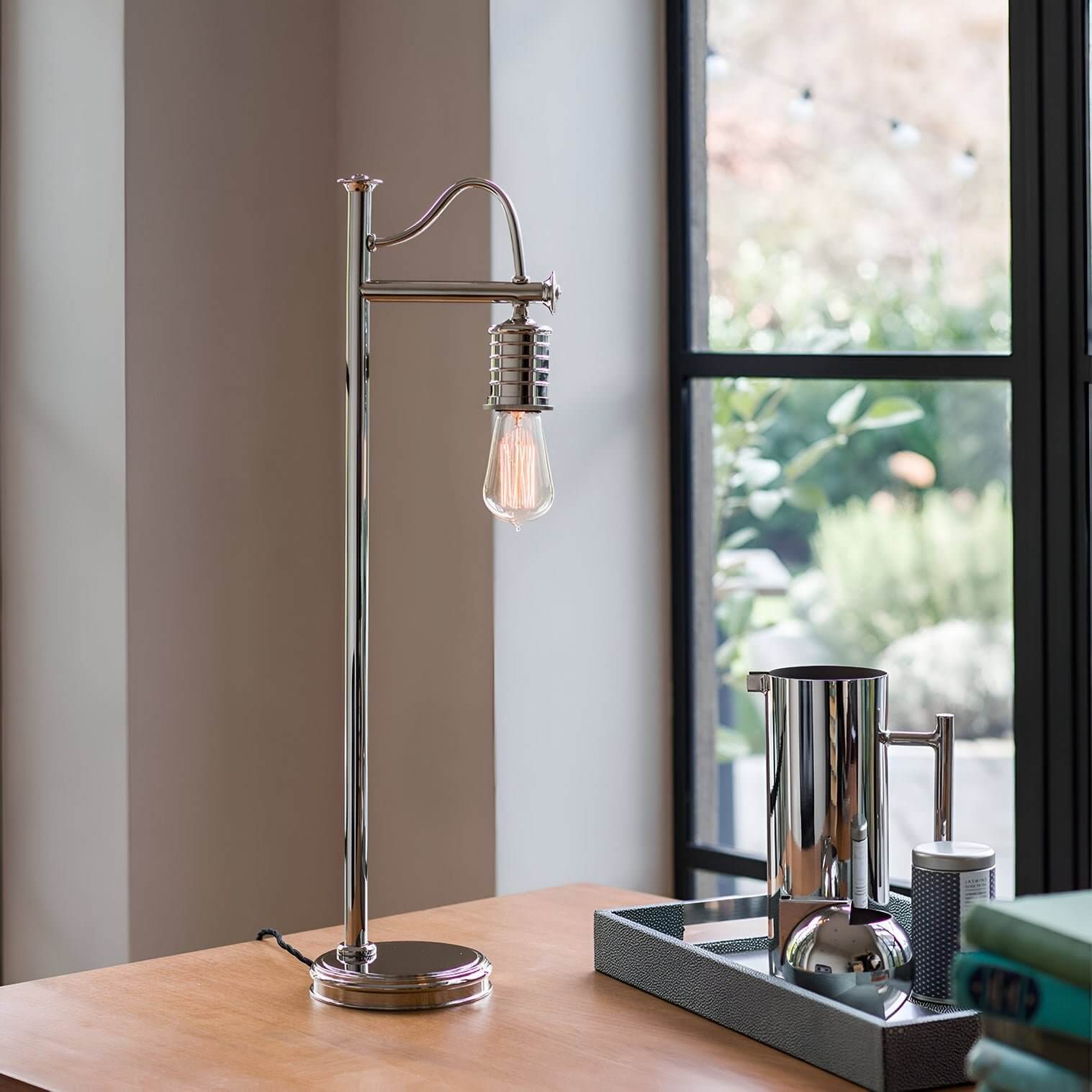 Slim chrome desk lamp with vintage bulb on a wooden table near a window, styled with modern metallic accessories.