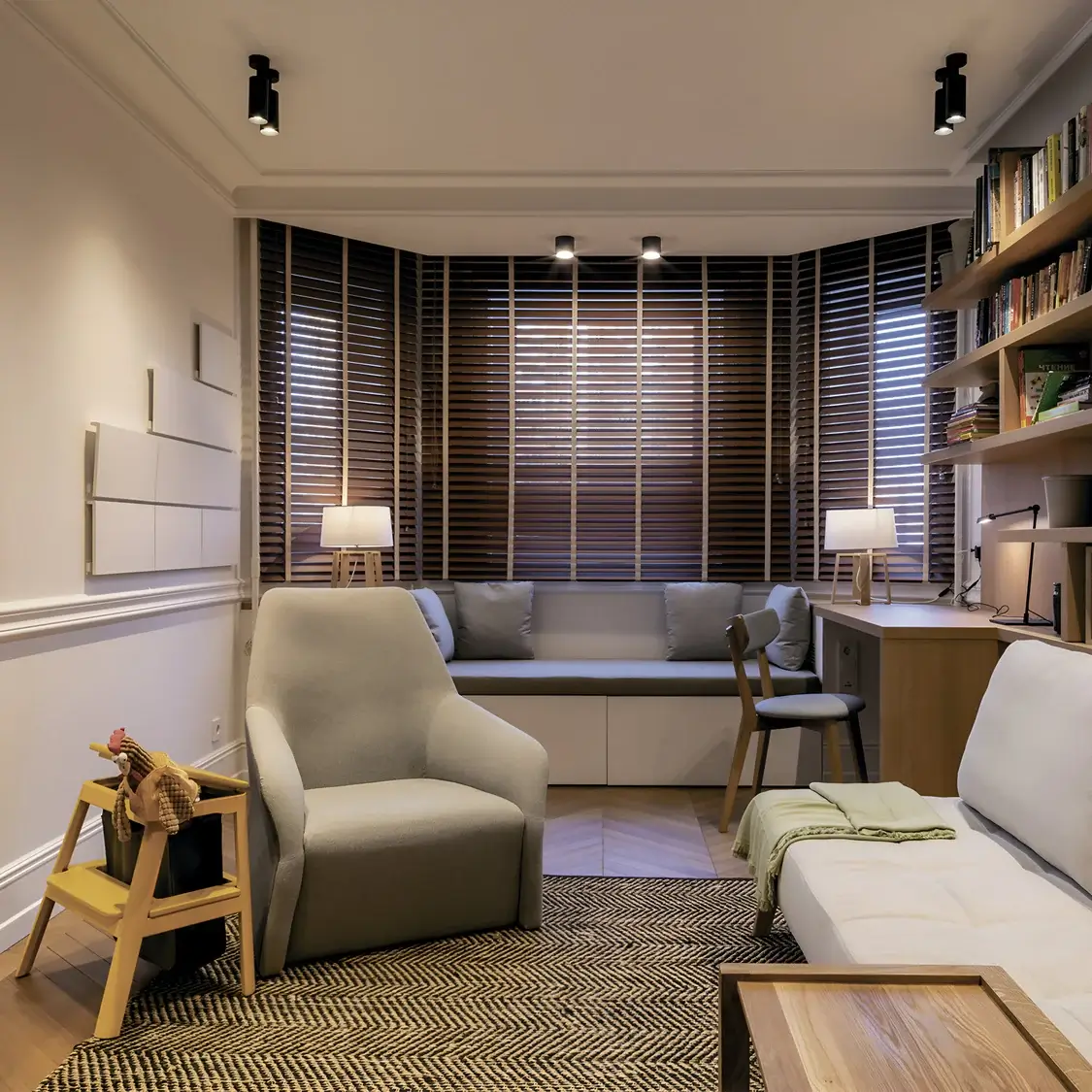 A compact living space with layered lighting, including ceiling-mounted spotlights and cozy table lamps. Wooden blinds and built-in bookshelves add texture and functionality, creating a serene reading or work environment.