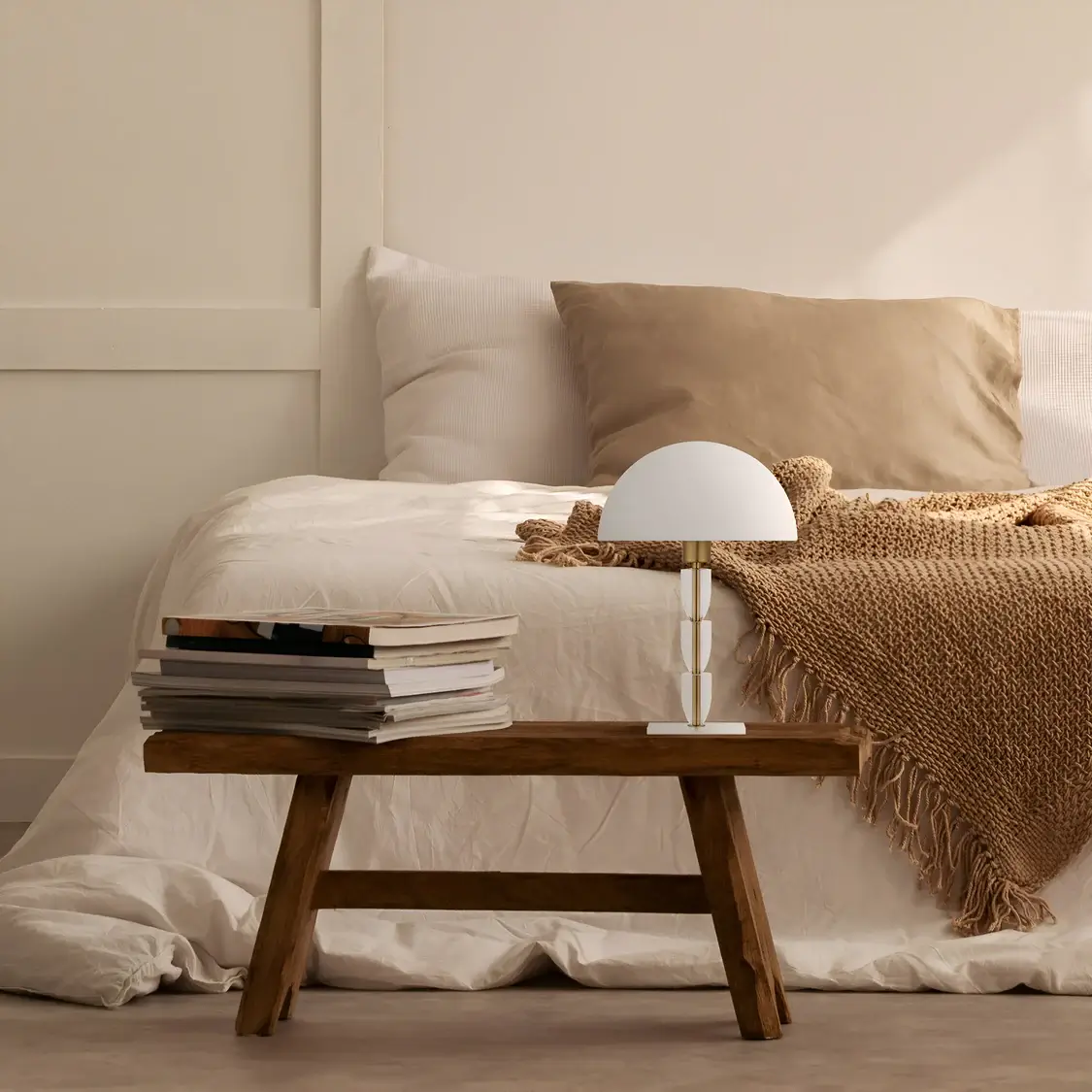 A rustic-inspired bedroom featuring a wooden bench used as a nightstand, topped with a stack of books and a table lamp with a white dome shade, surrounded by beige and neutral bedding and soft textures.