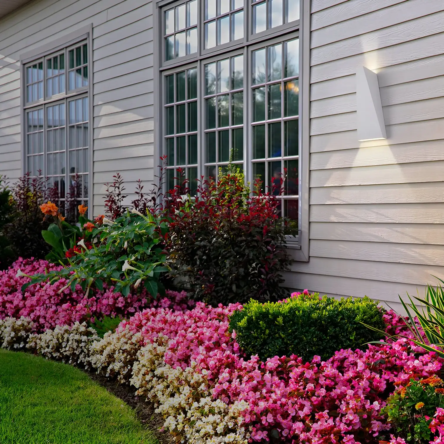 House exterior with bright white wall lights illuminating large windows and colorful flower beds along the garden path.