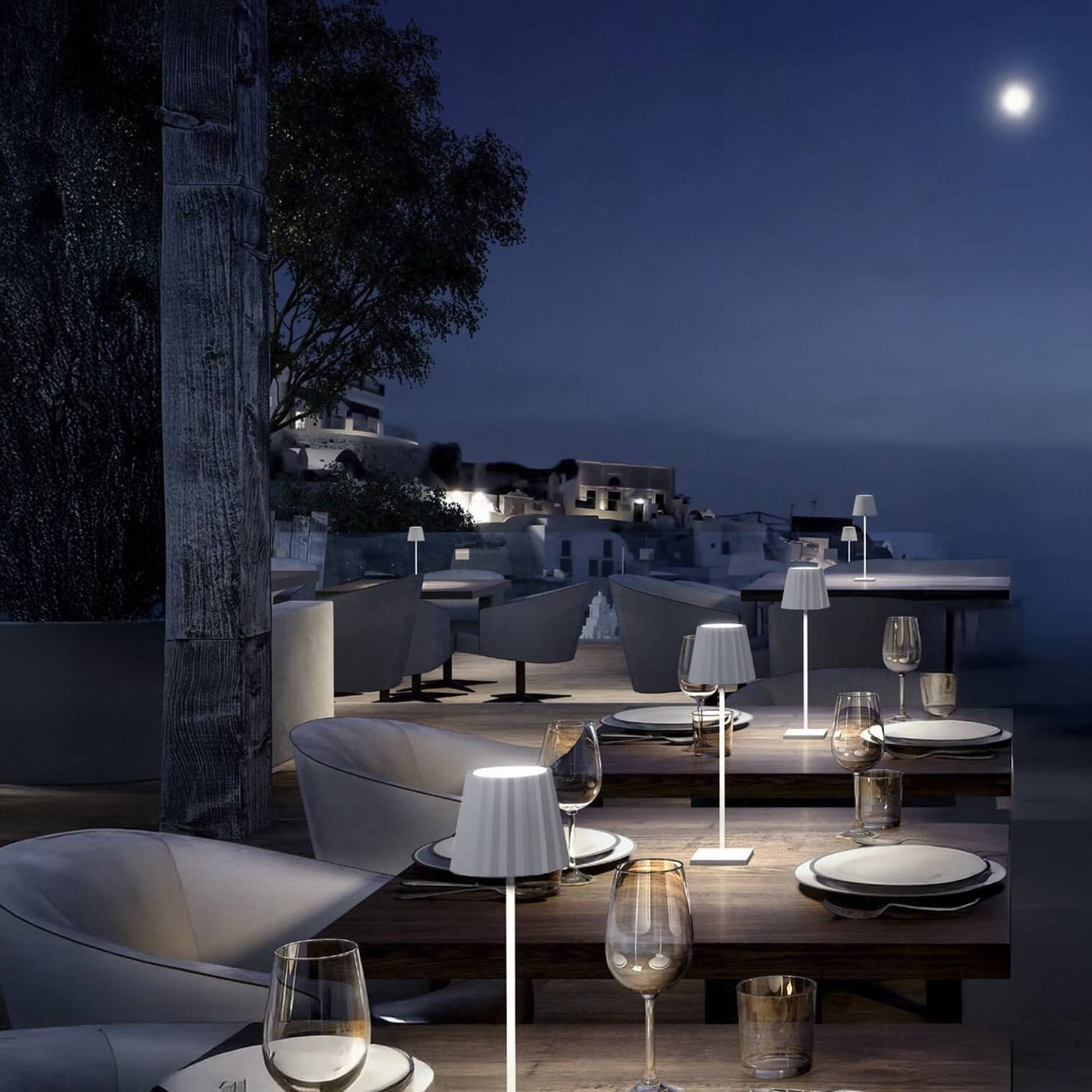 Sleek white cordless table lamps glowing on outdoor restaurant tables at night.