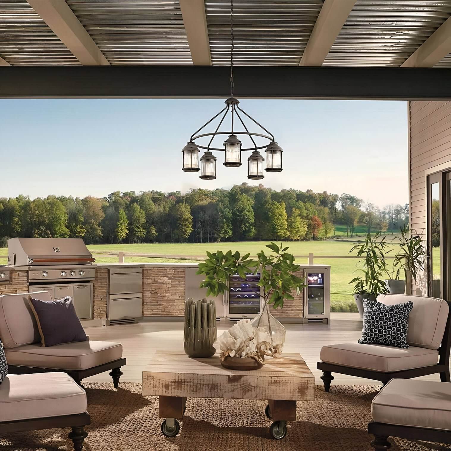 Open-air seating area with farmhouse chandelier, stone outdoor kitchen, and a view of a lush, tree-lined field.