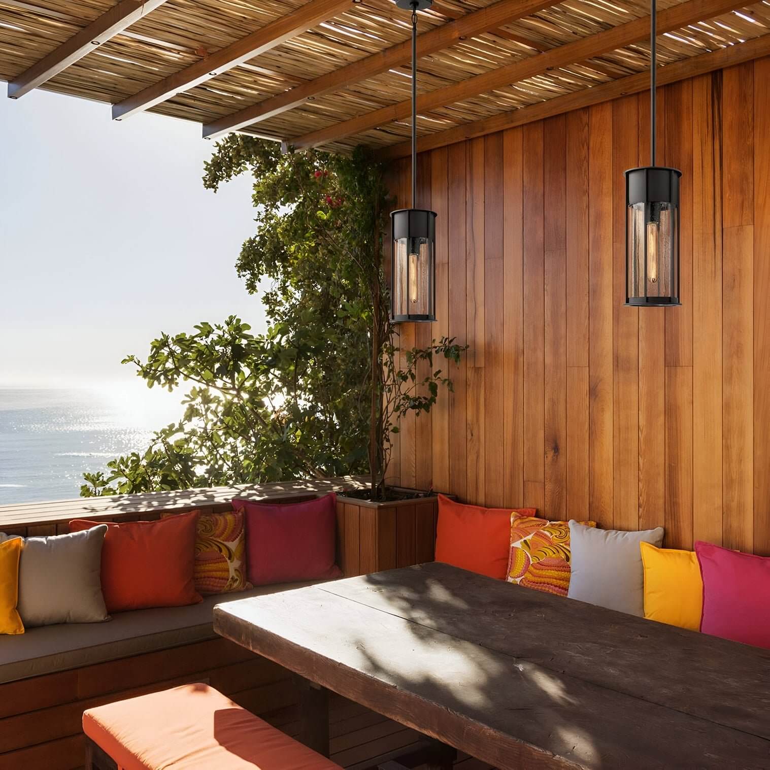 Wood-paneled patio with ocean view, bench seating, colorful pillows, and two cylindrical pendant lights hanging overhead.