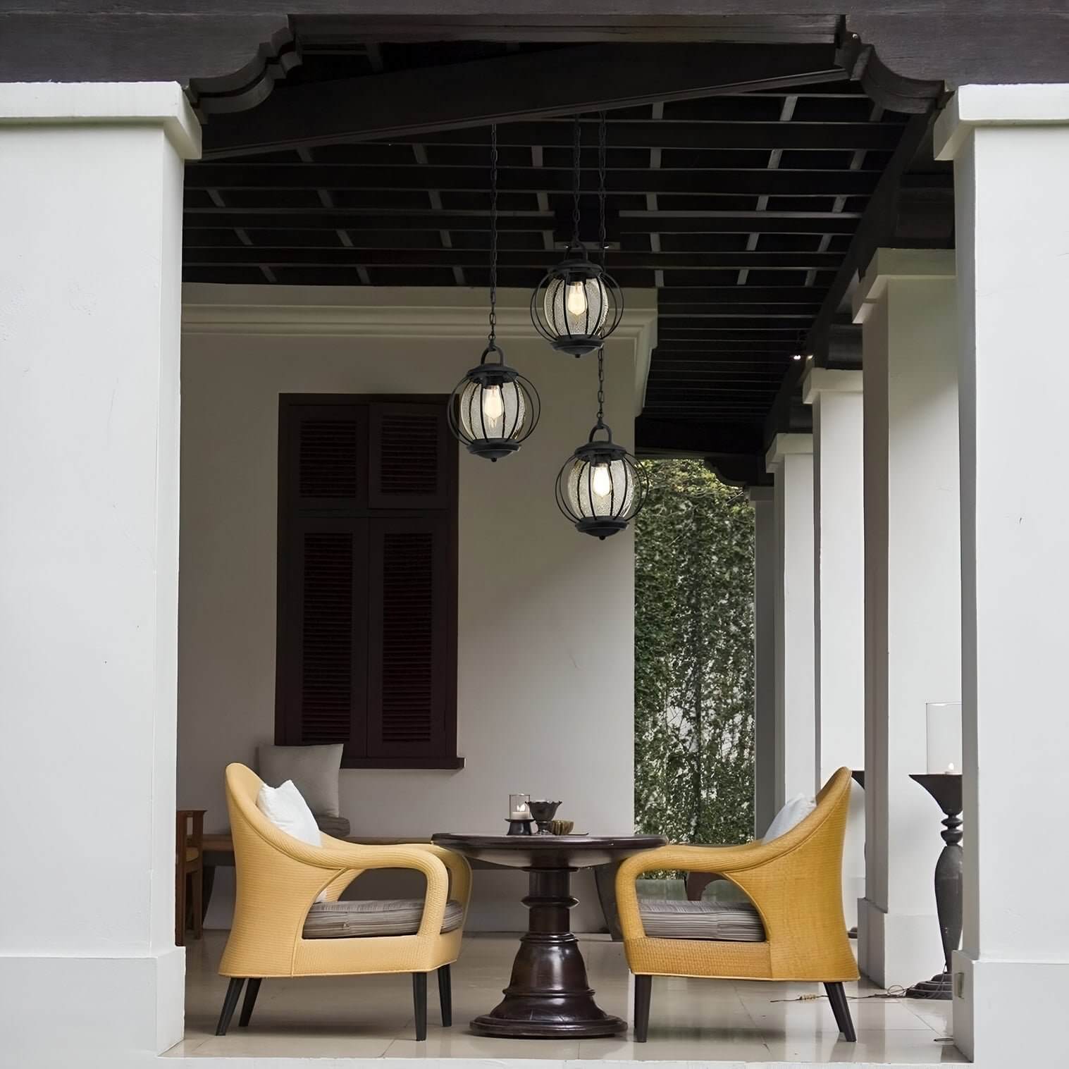 Covered patio with three black globe pendant lights, wicker chairs, and a small round wooden table in front of shuttered windows.